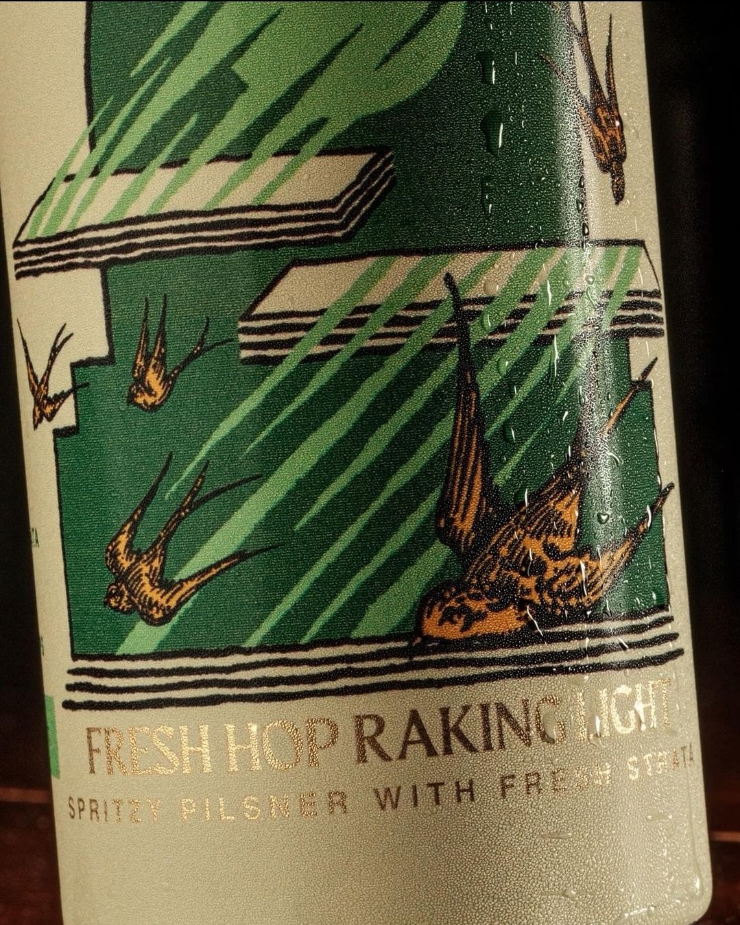 Close-up of a beer can label featuring a design with birds and abstract shapes in green and white. The text reads "Fresh Hop Raking Light, Spritzy Pilsner with Fresh Steath."