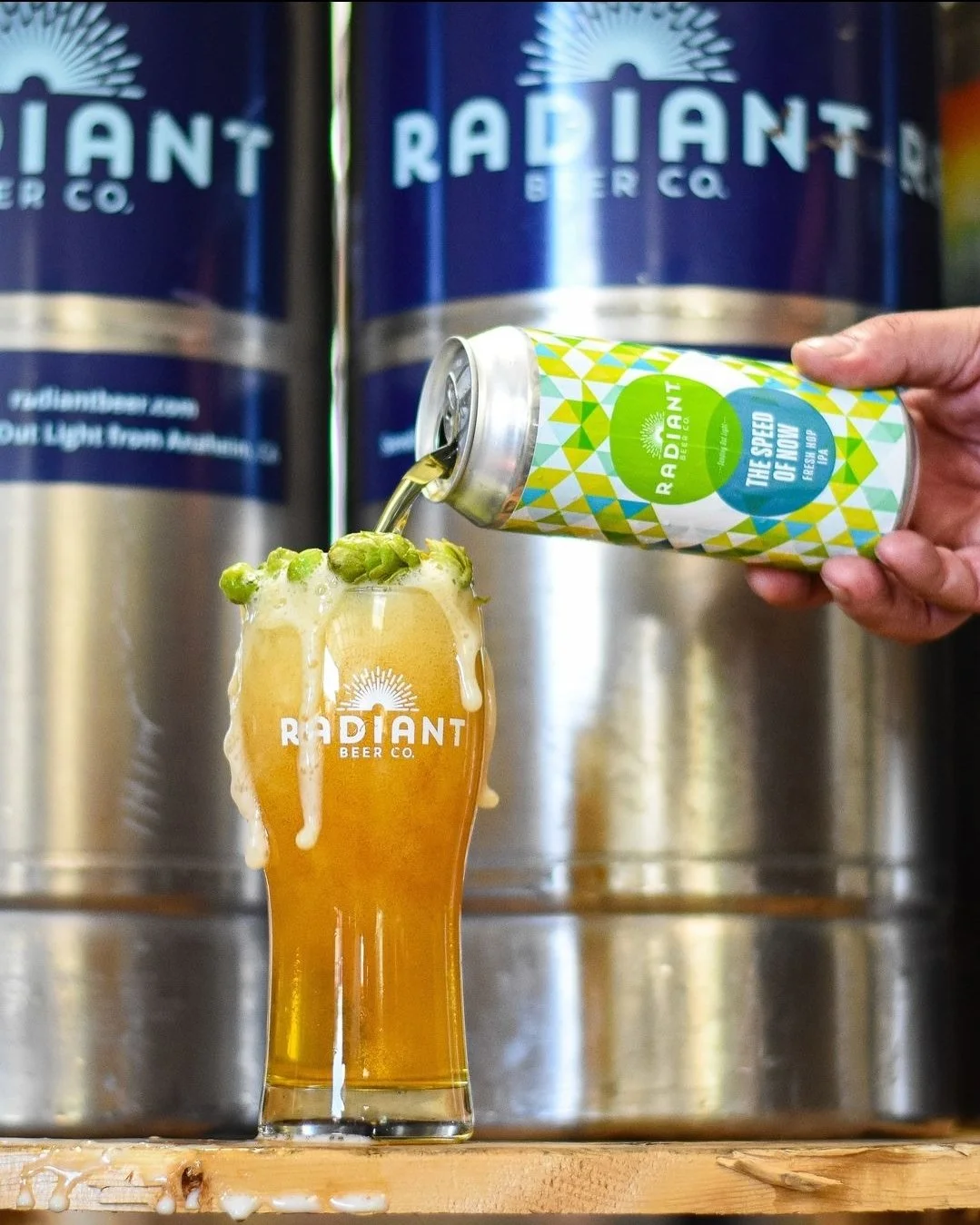 Radiant Beer Co. can pouring beer into branded glass with foam overflow, hops garnish, keg backdrop.