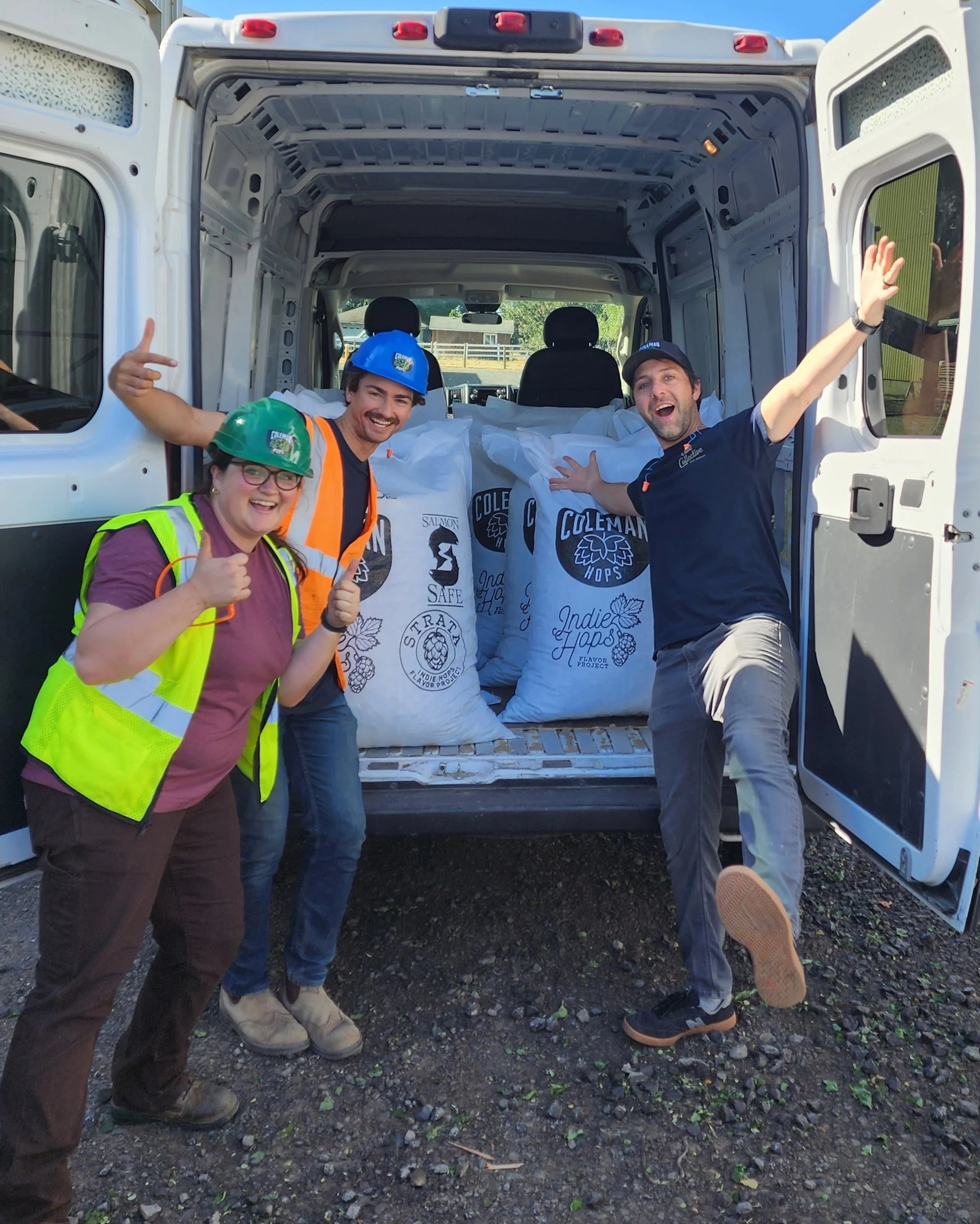 Three people, wearing safety vests and helmets, pose happily in front of a van loaded with bags labeled 'Coleman Hops' and 'Indie Hops.'
