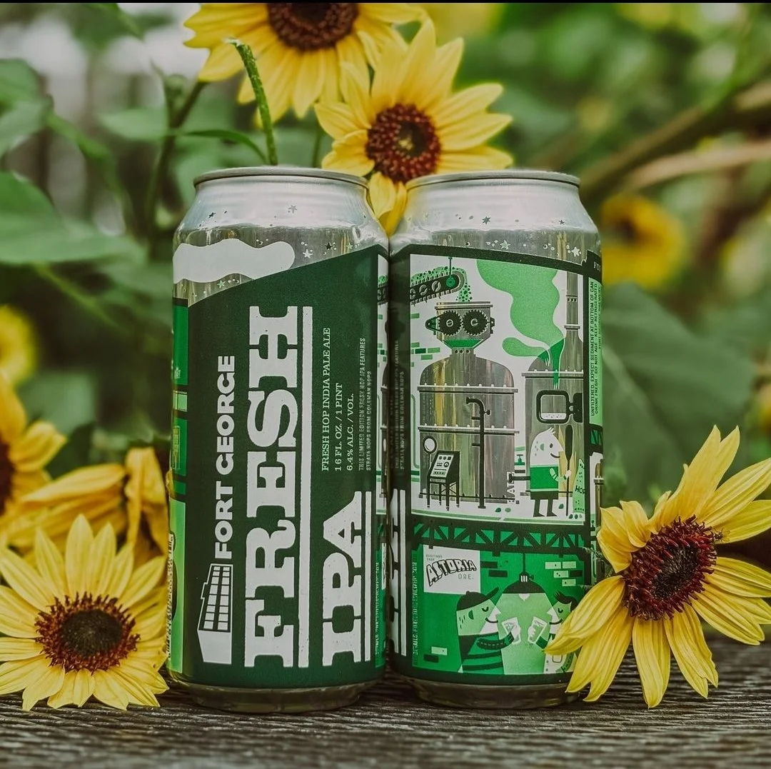 Two cans of Fort George Fresh IPA, surrounded by yellow sunflowers, with green foliage in the background. The cans feature detailed illustrations and text.