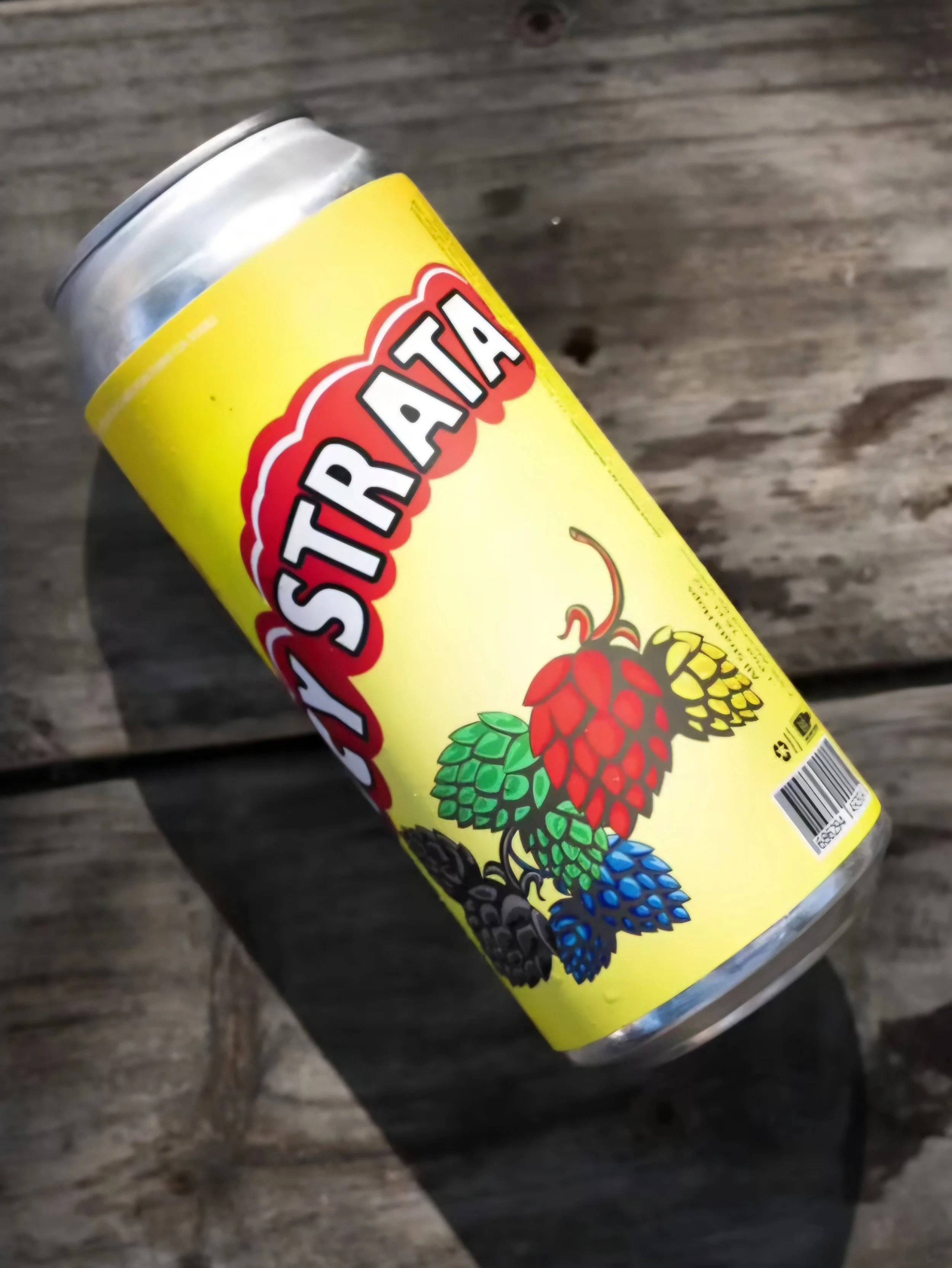 Yellow can of beer with 'Strata' label and colorful hop illustrations on a wooden surface.