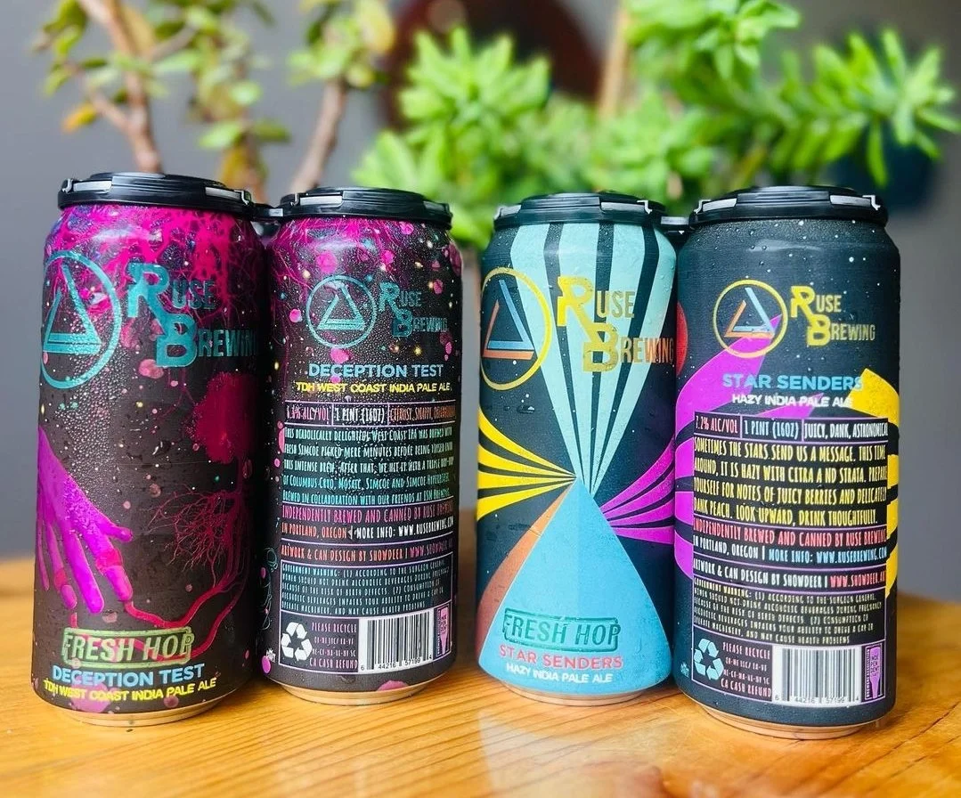 Four colorful beer cans from Ruse Brewing, featuring designs for 'Deception Test' and 'Star Senders' IPAs. The cans display vibrant graphics with abstract art and beer details, placed on a wooden surface with plants in the background.