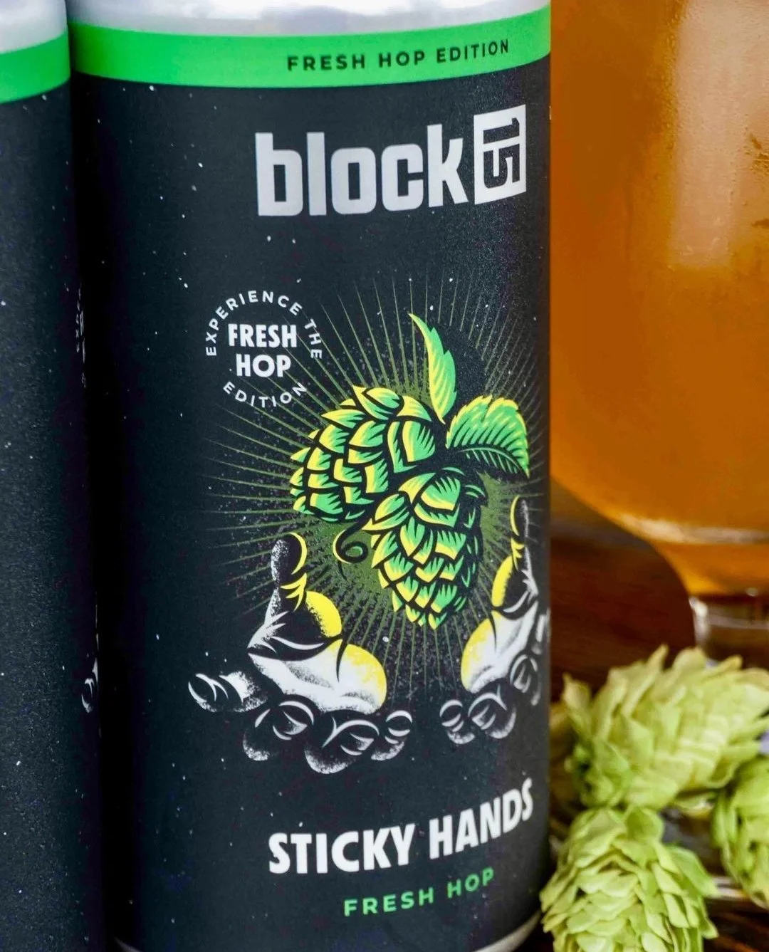 Can of "block 15 Sticky Hands Fresh Hop" beer with hops graphic, glass of beer, and fresh hop cones on a wooden surface.