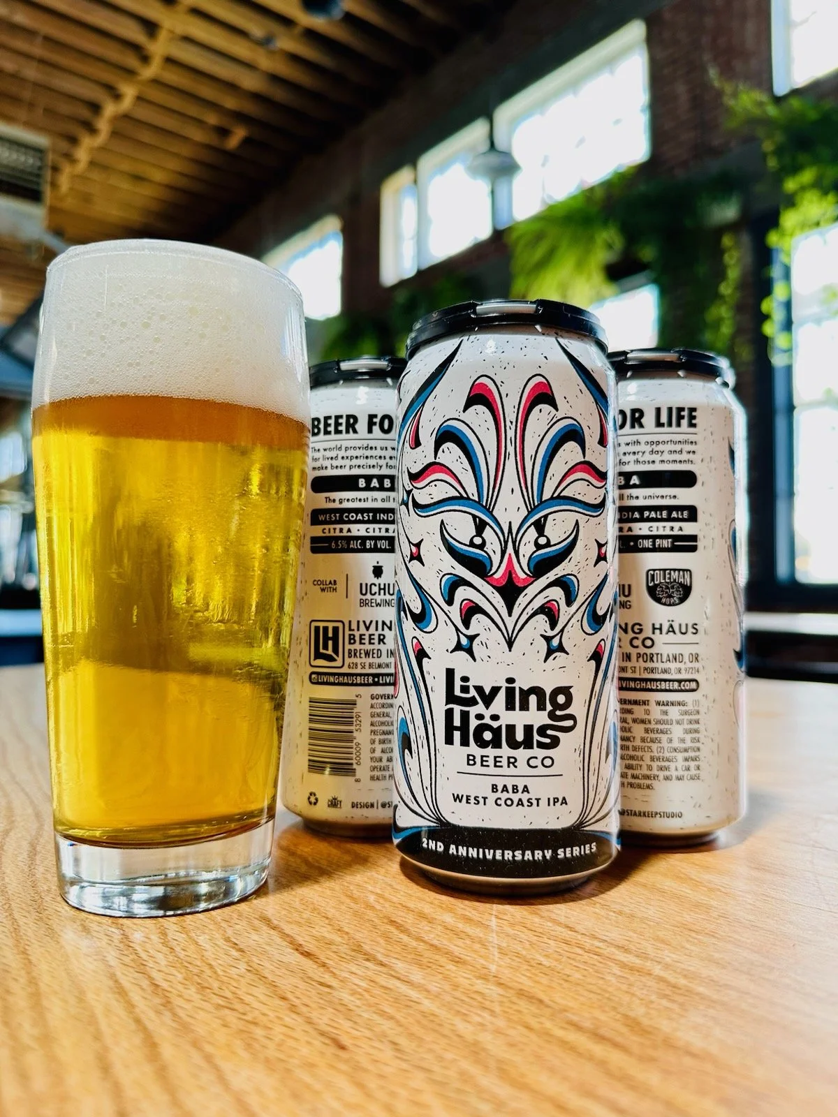 A can of Living Haus Beer Co.'s 2nd Anniversary Series, BABA West Coast IPA, next to a pint glass filled with beer, on a wooden table in a brewery setting.