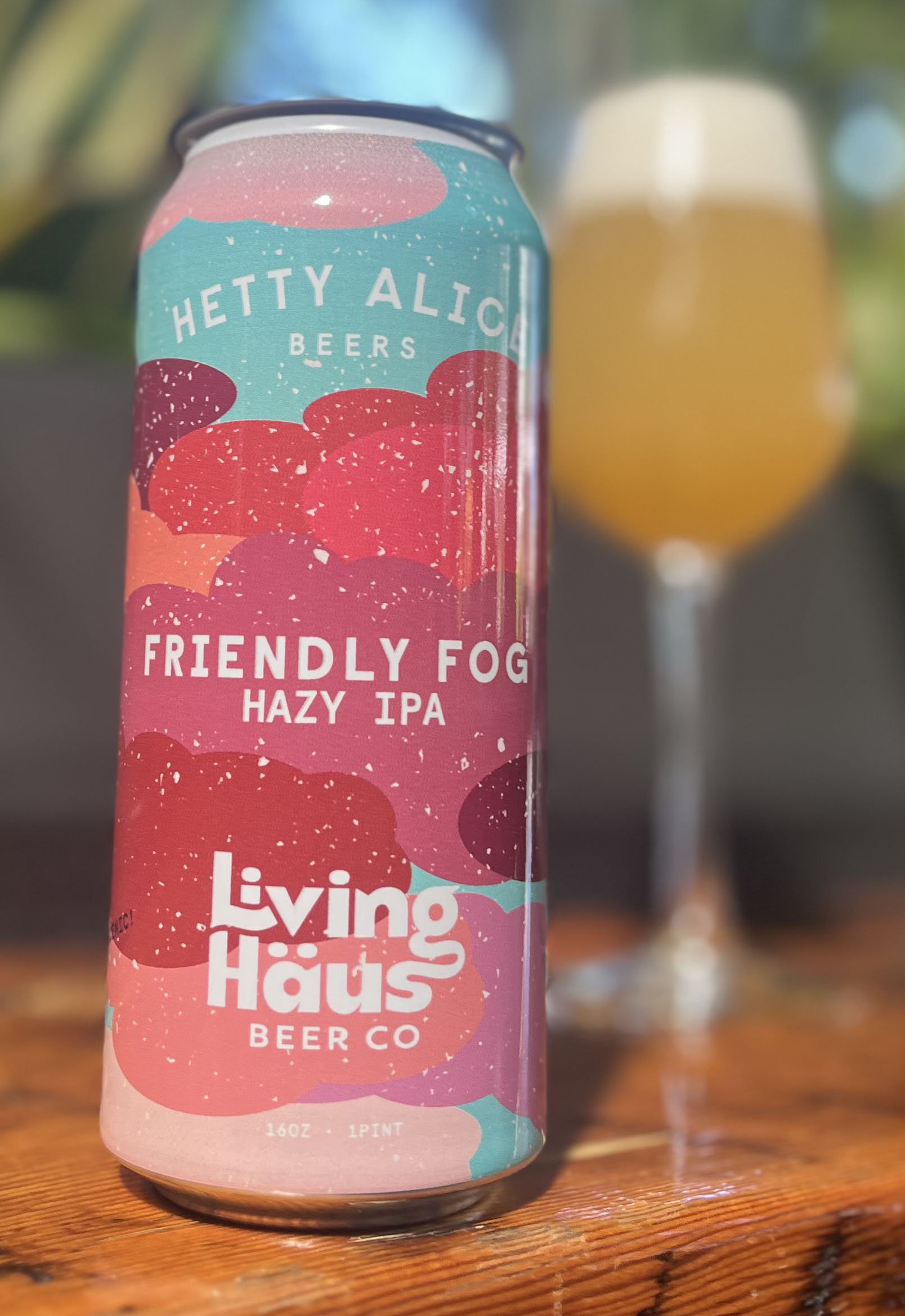 A colorful can of Hetty Alice Beers Friendly Fog Hazy IPA in front of a glass of beer, placed on a wooden surface.