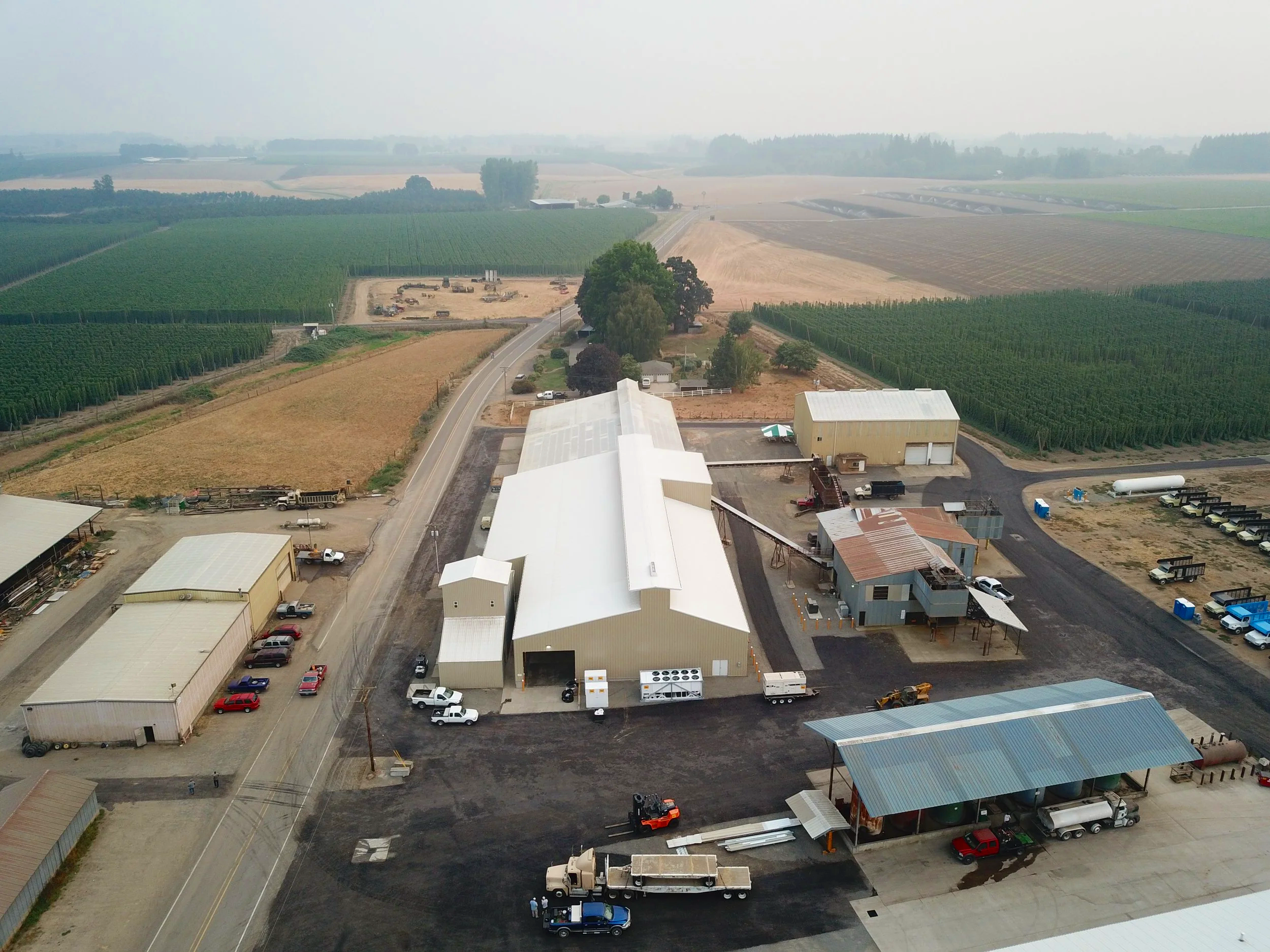 Aerial view of a farm with large white metal buildings, surrounded by cultivated fields, farm equipment, and parked vehicles.