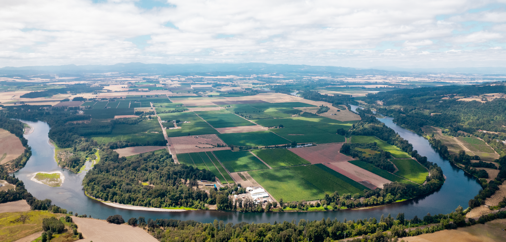 Aerial view of a winding river running through farmland with fields, trees, and a few buildings under a partly cloudy sky.