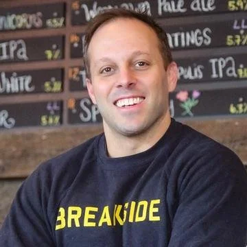 A smiling man with short hair wearing a black sweatshirt with yellow text, standing in front of a chalkboard menu at a brewery or bar.