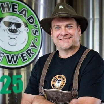 A man wearing a brown hat and black T-shirt with a logo, standing in front of a brewery sign that reads "Fat Head's Brewery" and displays the number 25.