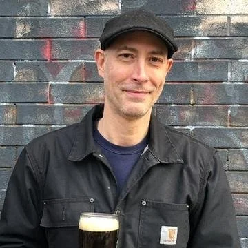 Man wearing black jacket and baseball cap standing in front of brick wall, holding a glass of dark beer.