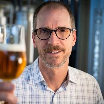 A middle-aged man with glasses and a beard holding up a glass of beer in a social setting.