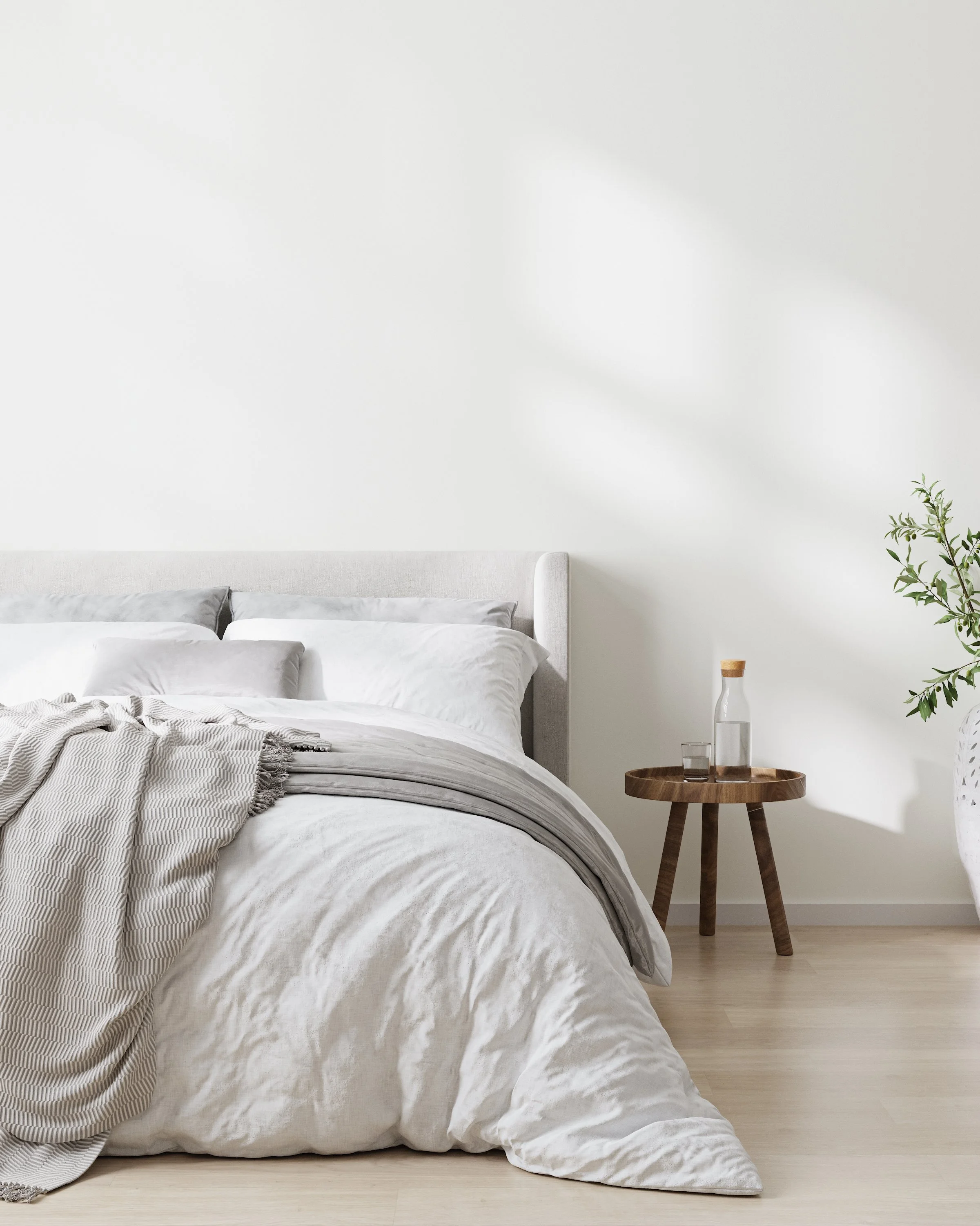 A neatly made bed with white and light gray bedding, a beige upholstered headboard, and a small wooden side table with a glass water carafe and a glass, adjacent to a potted plant in a white decorative pot, all set against a plain white wall.