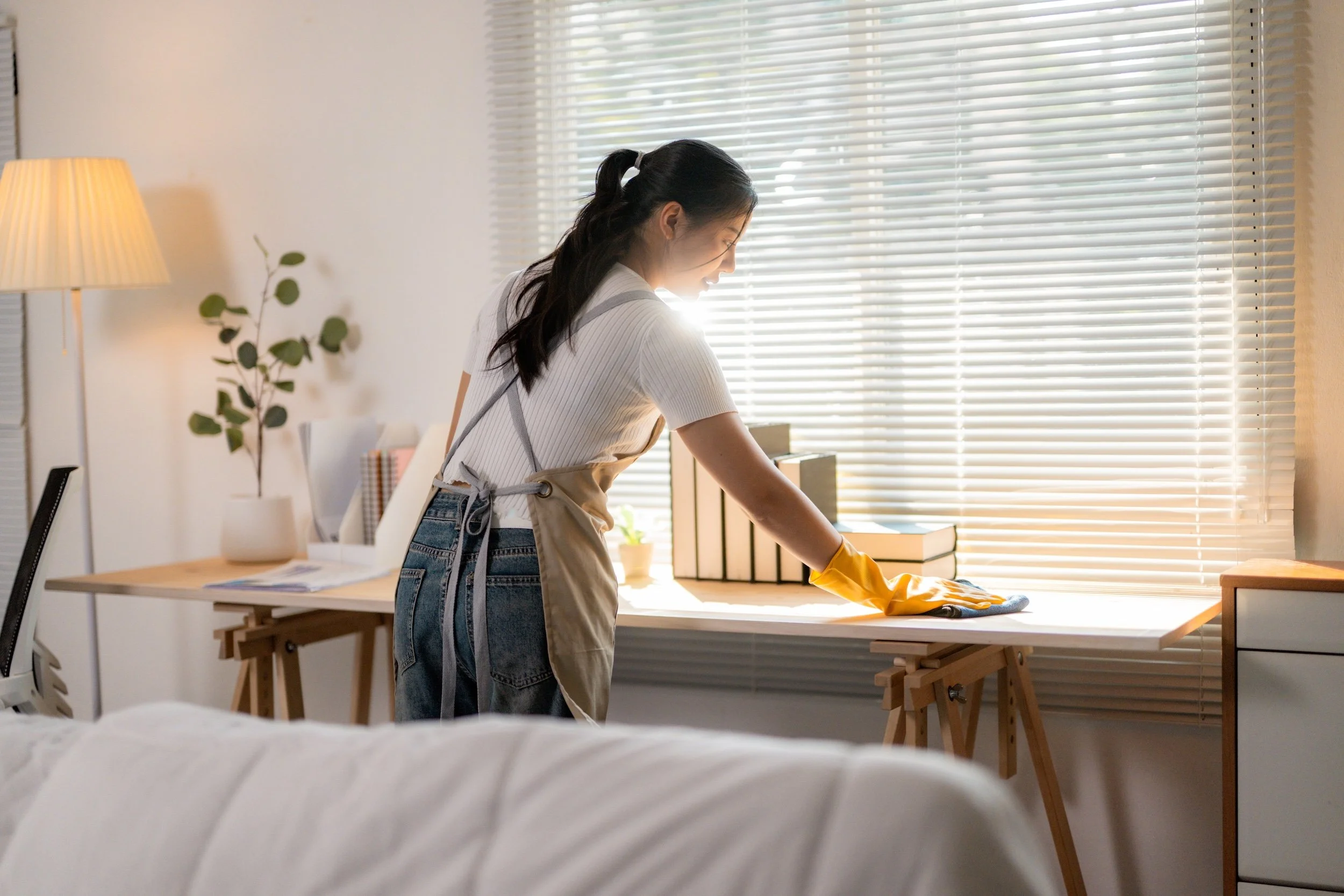 A woman cleaning a bright bedroom workspace, wiping a desk near a sunlit window with yellow gloves and an apron.