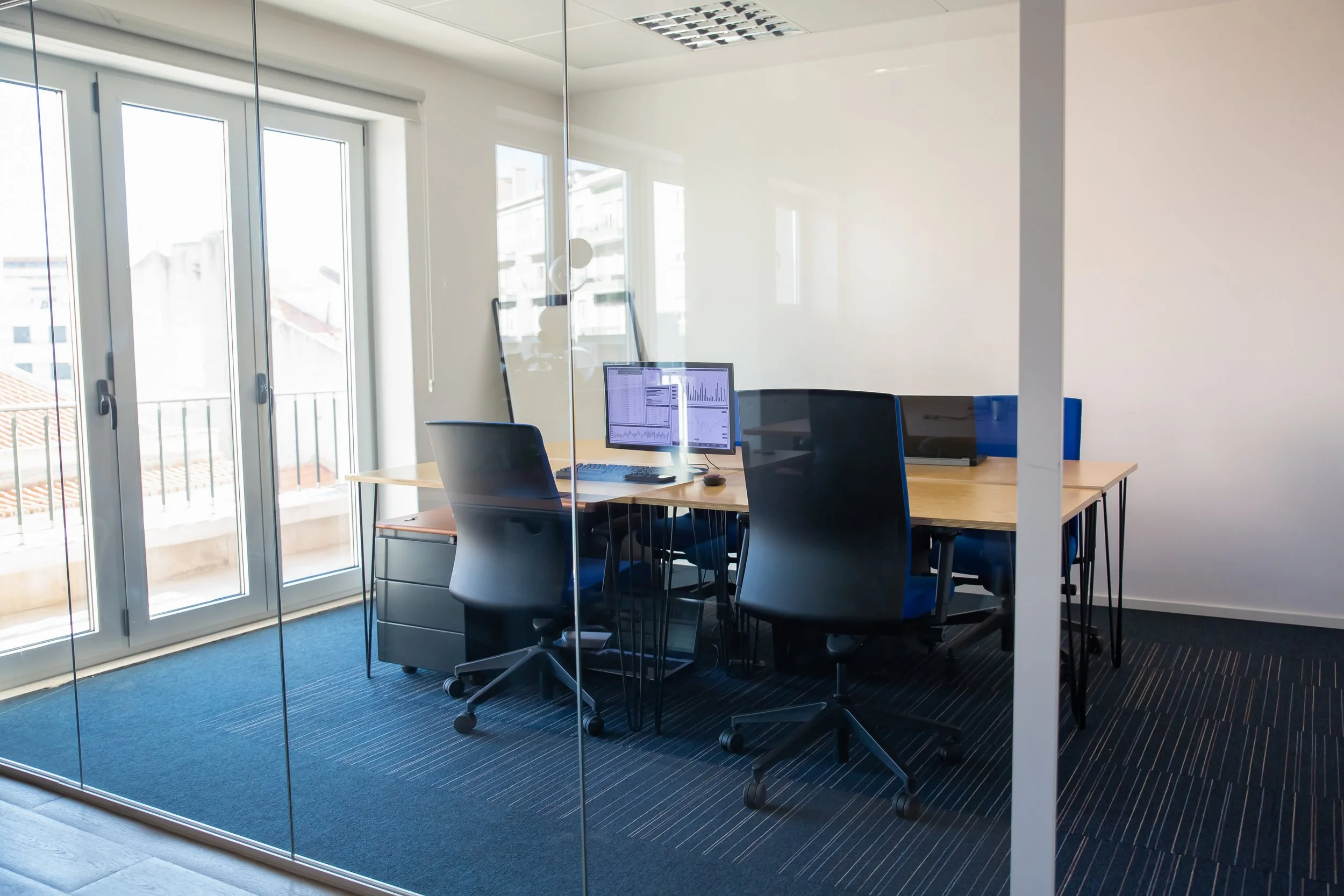 An empty modern office conference room with glass walls, a wooden table, black chairs, computer monitors, and windows letting in sunlight.