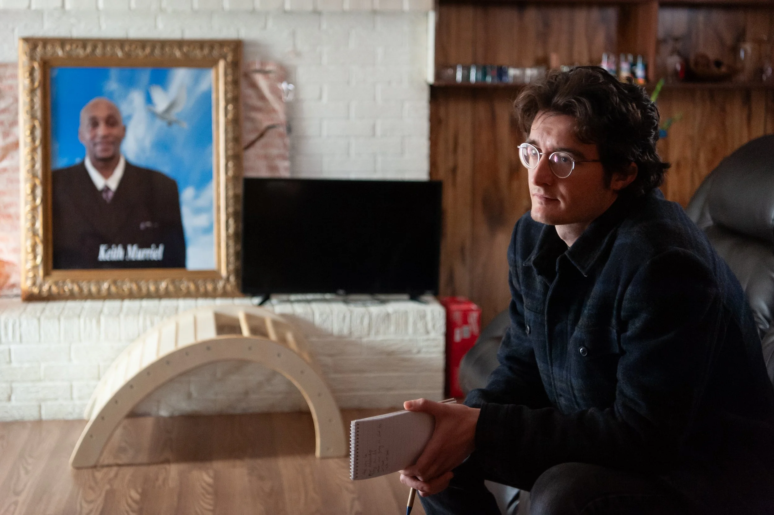 A man with curly hair and glasses sitting on a black leather couch, holding a notepad and pen, in a living room with a framed portrait of a man named Keith Muriel on the wall behind him.