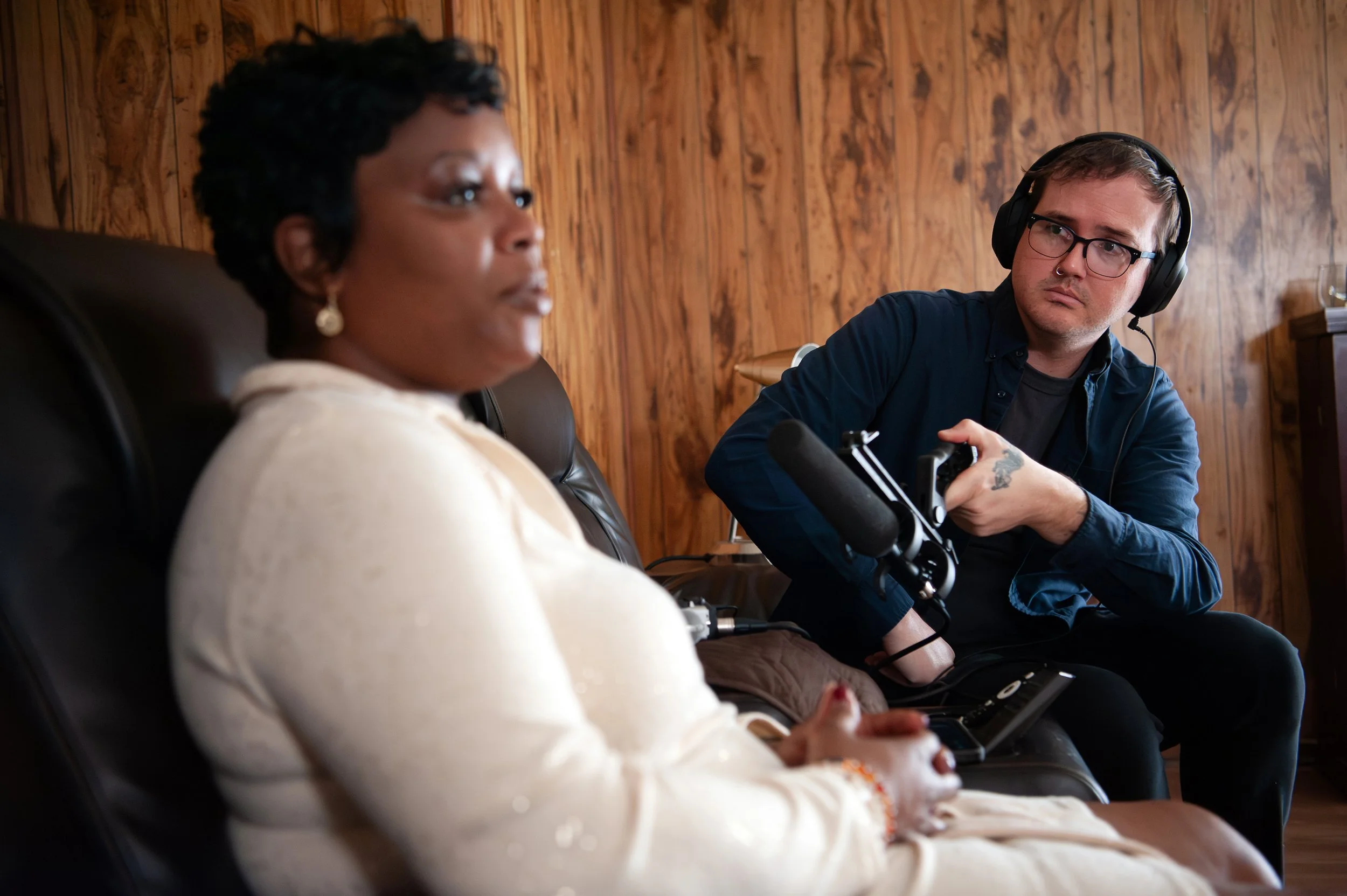 A woman with dark curly hair and earrings sitting on a leather chair with a man wearing headphones, glasses, and a blue shirt, holding a microphone, in a room with wooden walls.