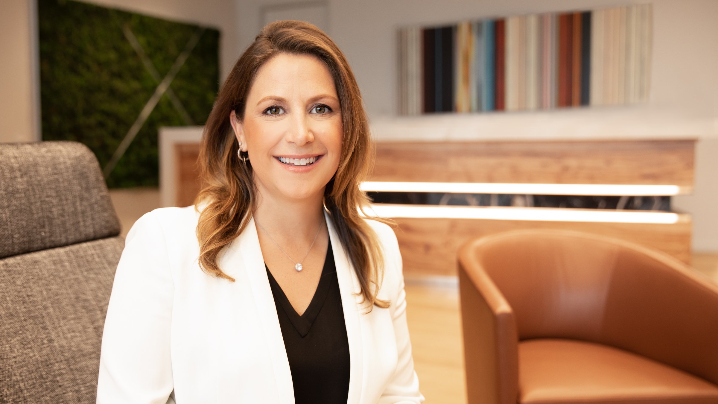 A professional woman with shoulder-length brown hair, smiling, sitting in a modern office space with a bookshelf and framed art in the background.