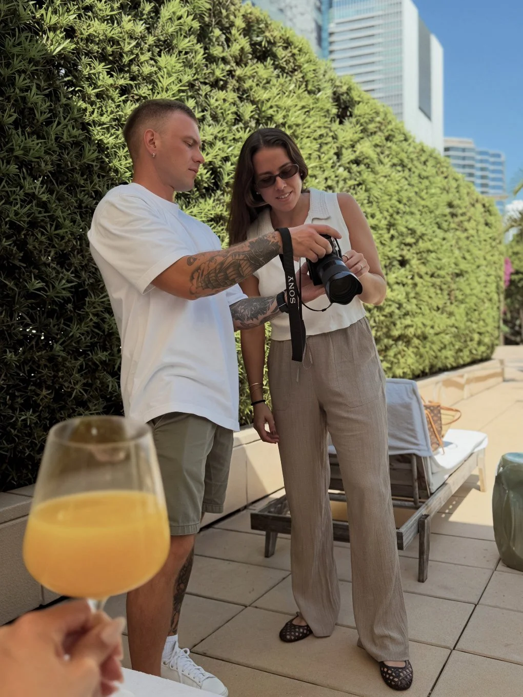Two people standing outdoors beside tall bushes, looking at a camera the man is holding. The woman is wearing sunglasses and tan pants. The man has tattoos on his arms and is wearing a white t-shirt and shorts. In the foreground, a hand is holding a glass of orange juice. There are modern buildings and a blue sky in the background.