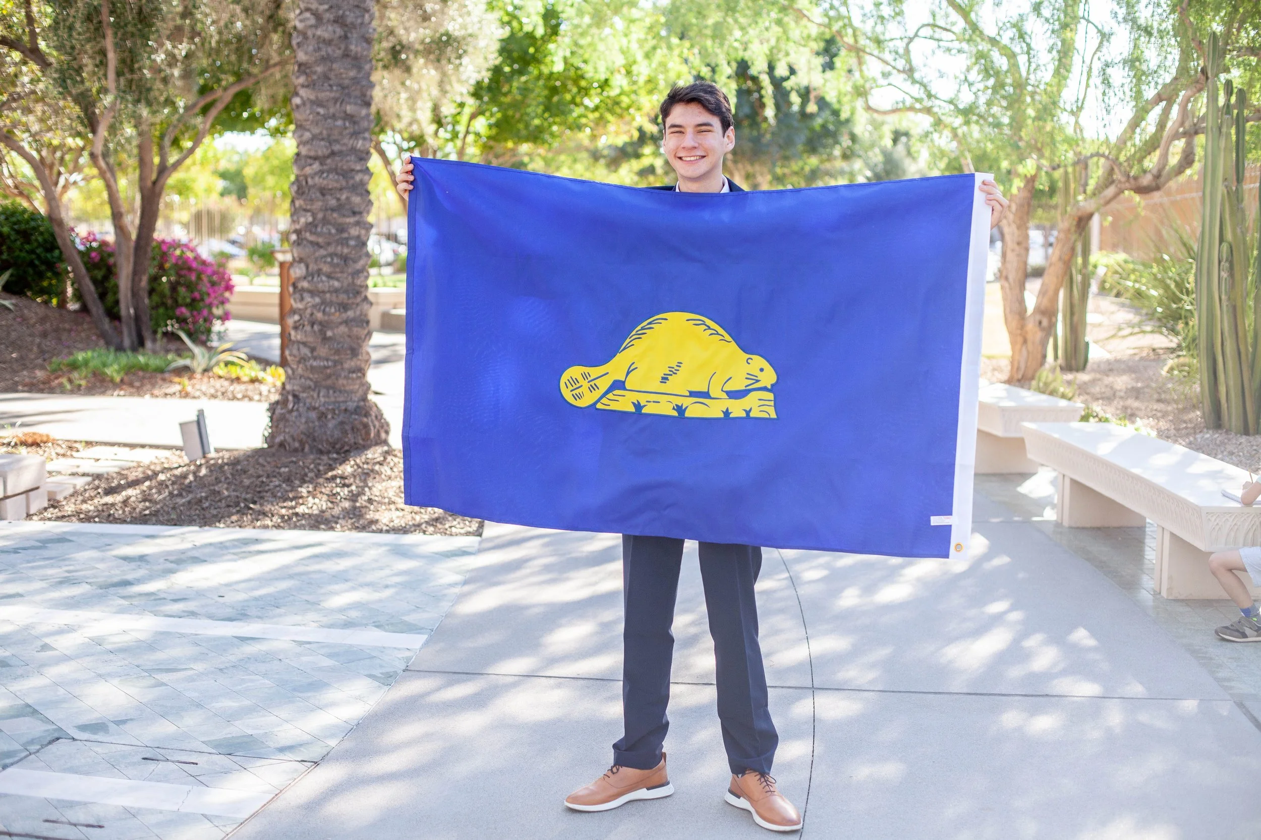A young man wearing a suit and tan shoes holding a Louisiana state flag outdoors with trees and plants in the background.
