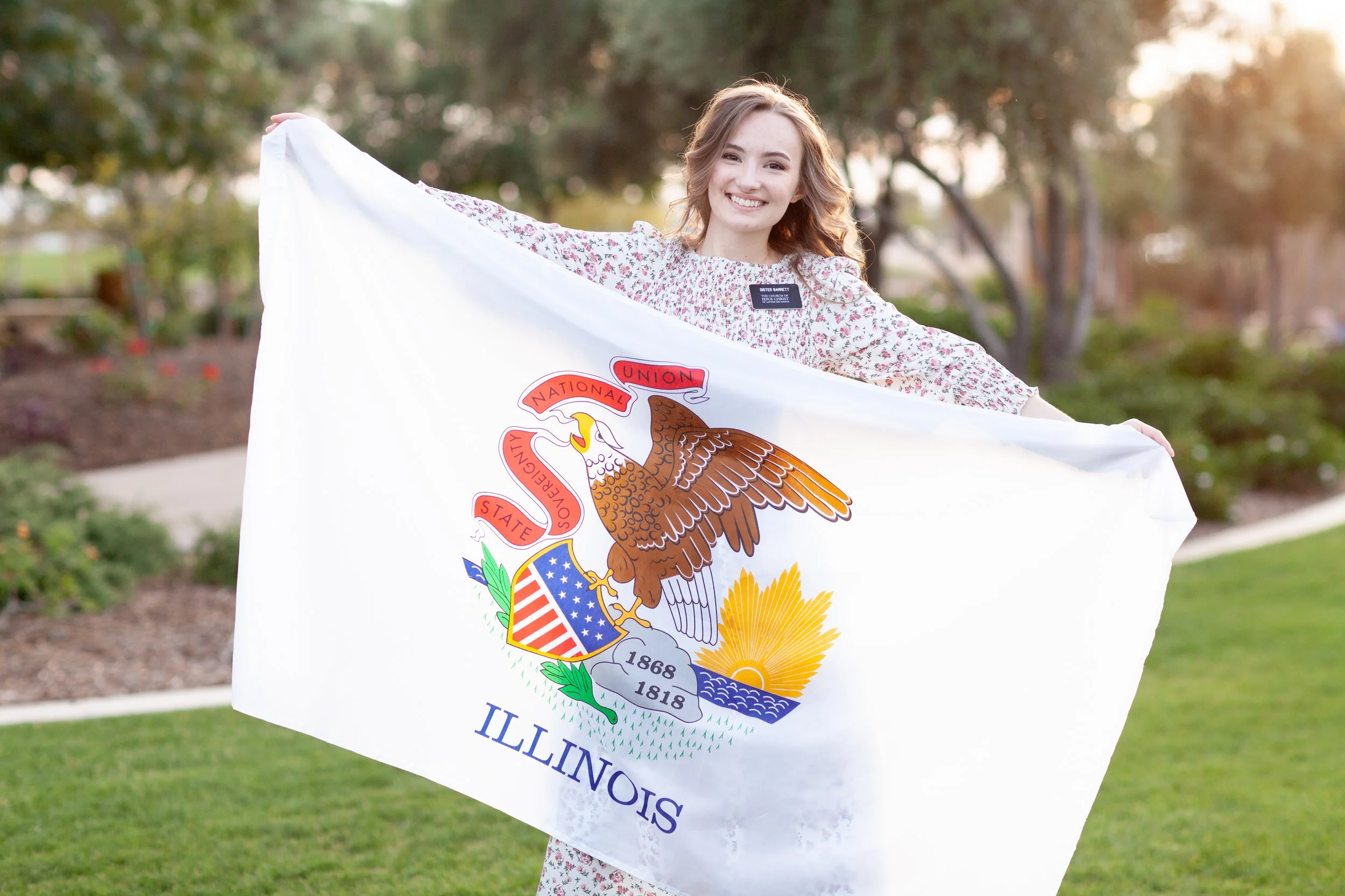 A young woman standing outdoors, smiling, holding a large white flag with the Illinois state seal on it, in a park with trees and grass, during the daytime.