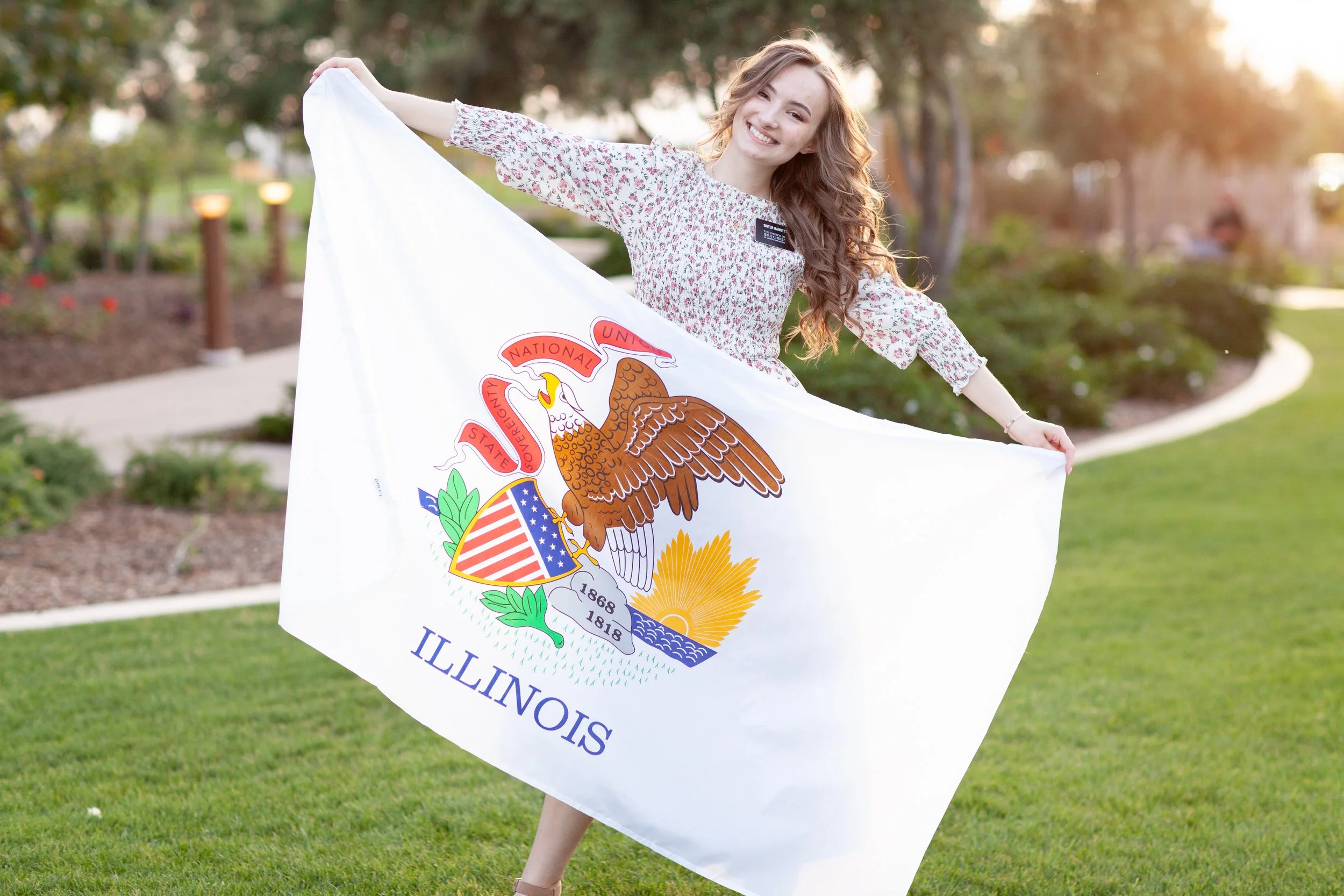 A young woman with long, curly brown hair smiling and holding the California state flag outdoors in a park during the daytime.