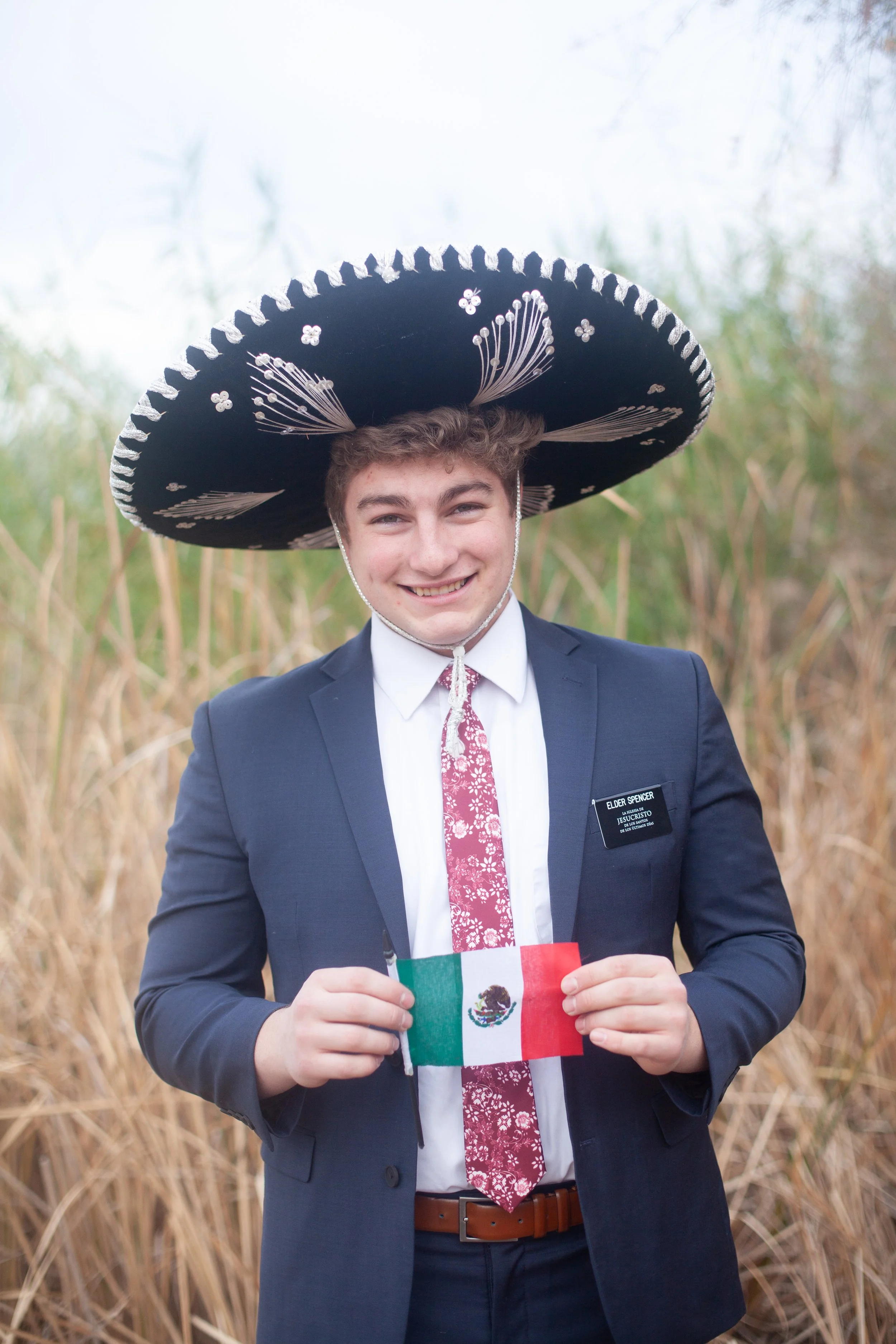A young man dressed in a suit and tie, smiling, wearing a large traditional Mexican sombrero, holding a small Mexican flag, standing outdoors among tall grass.