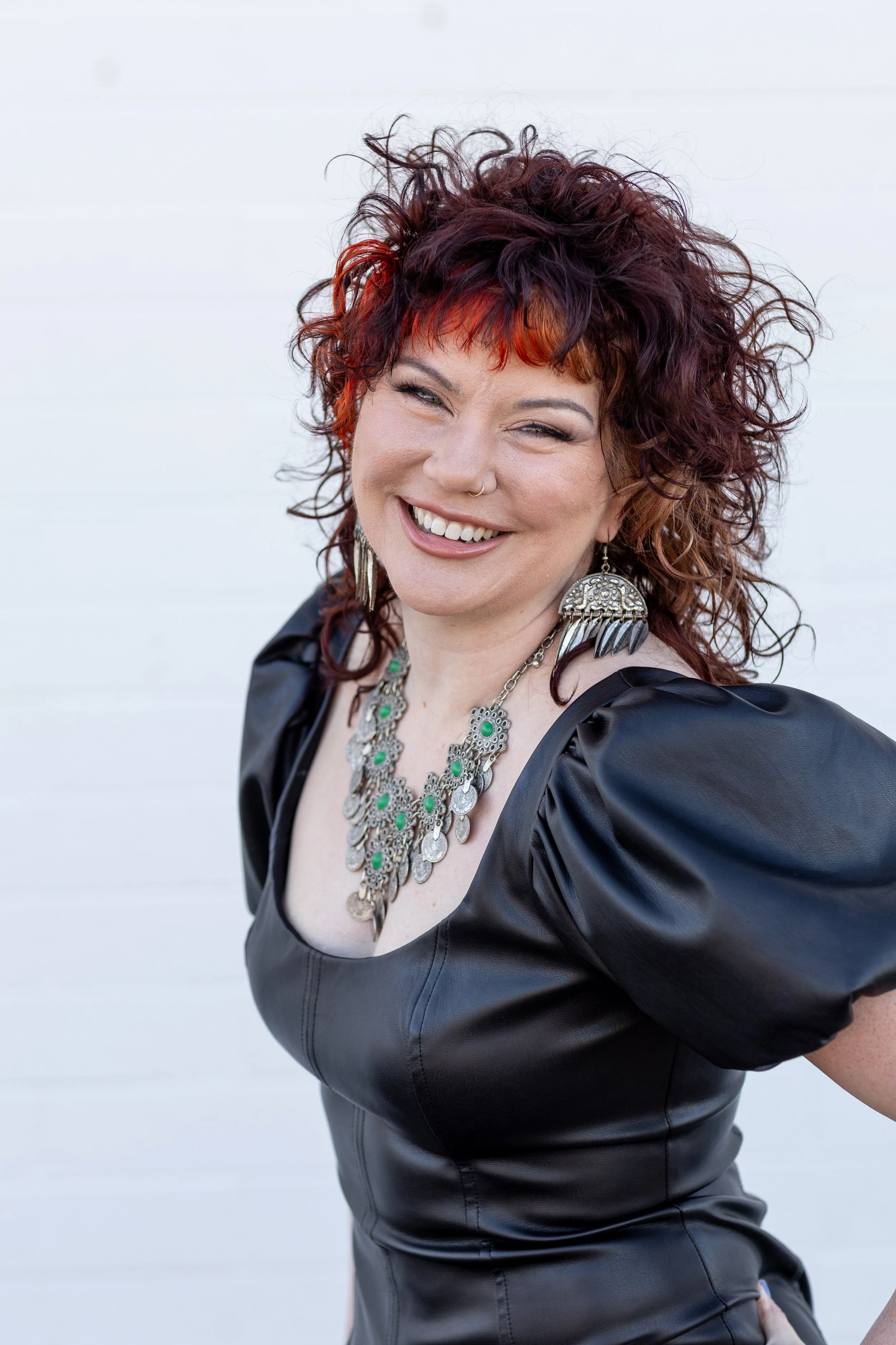 A woman with styled, curly hair with orange highlights, wearing a black leather dress and large ornate necklace, smiling against a white background.