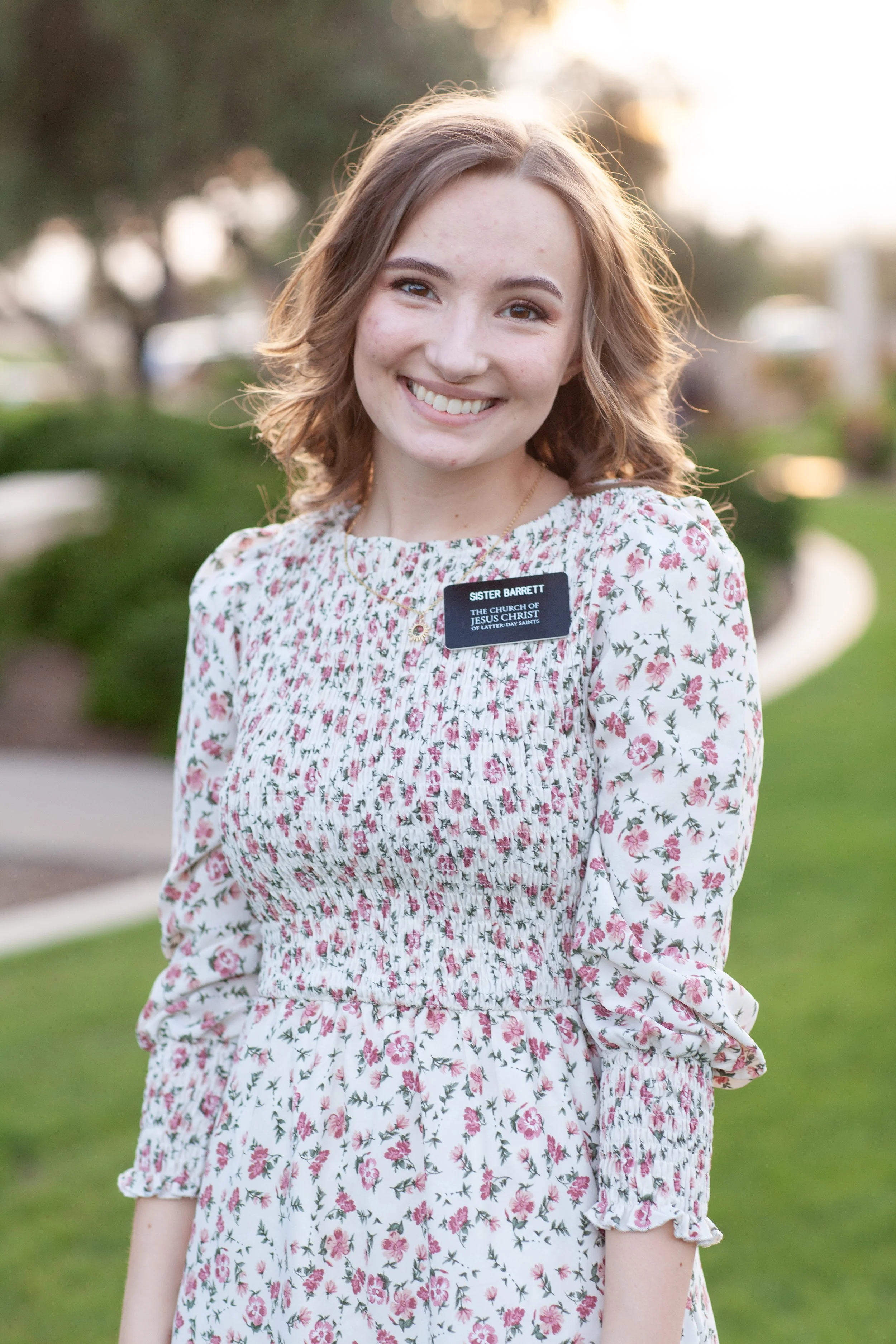 A young woman with light brown hair smiling outdoors in a floral dress with ruffled sleeves, standing in front of a blurred background of trees and grass during golden hour.