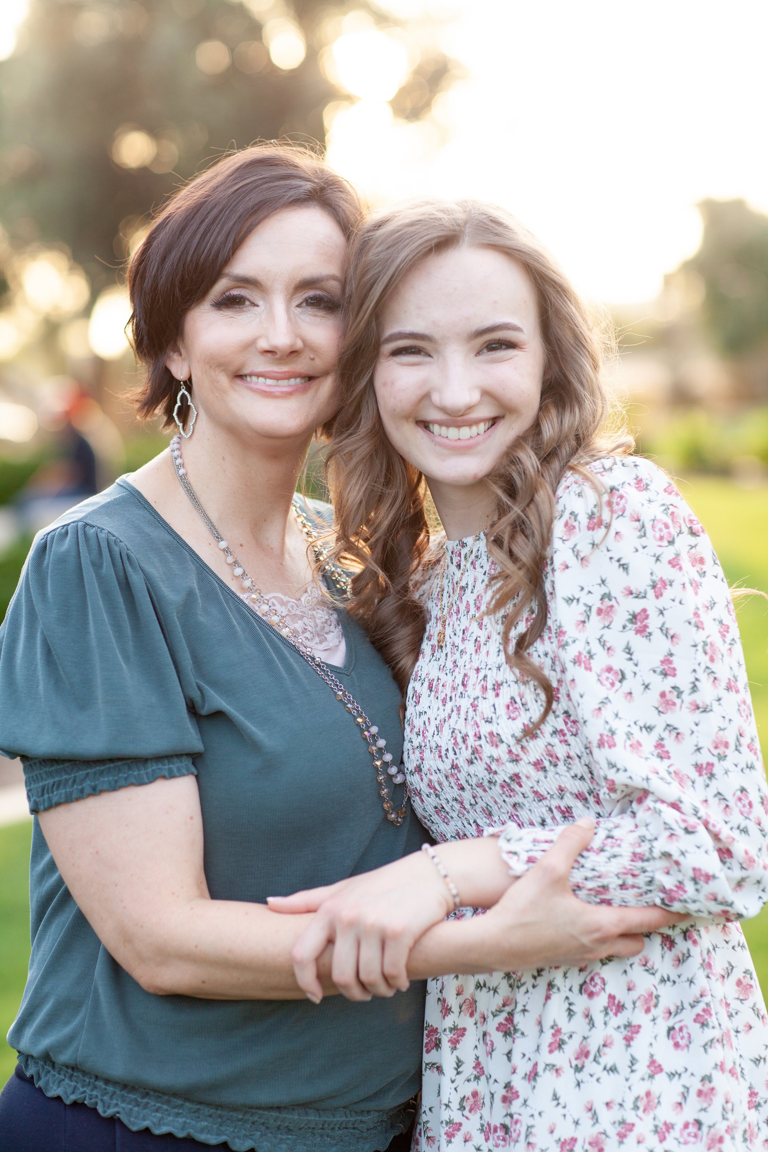 A woman and a girl smiling together outdoors at sunset, with the woman wearing a teal blouse and jewelry, and the girl in a floral dress.