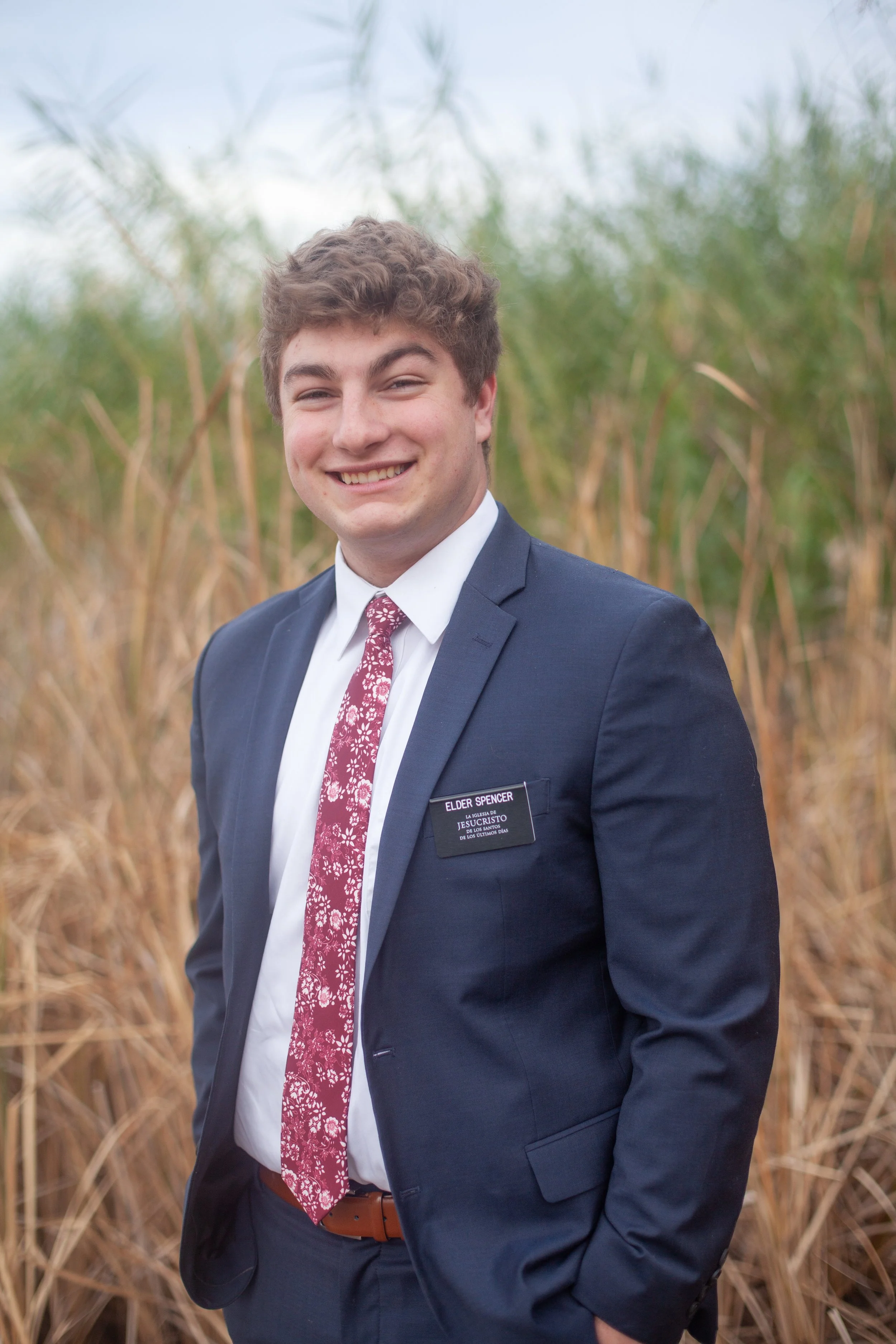 Young man in a navy suit with a patterned maroon tie, white shirt, and a name tag standing outside in a field of tall, dry grass or reeds, smiling at the camera.