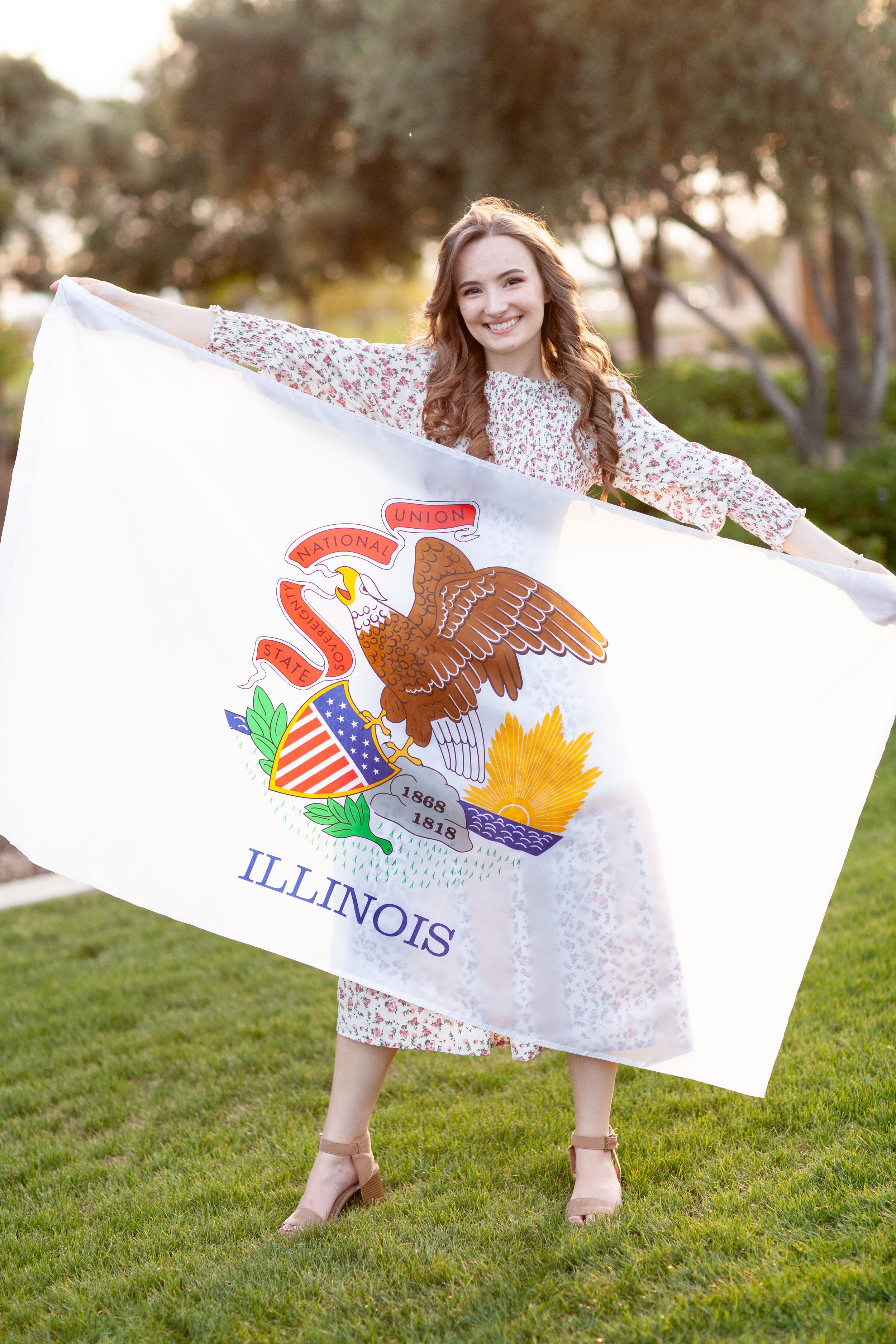 A young woman with long, wavy light brown hair, smiling and holding an Illinois state flag, standing outdoors on a grassy field with trees in the background during sunset.