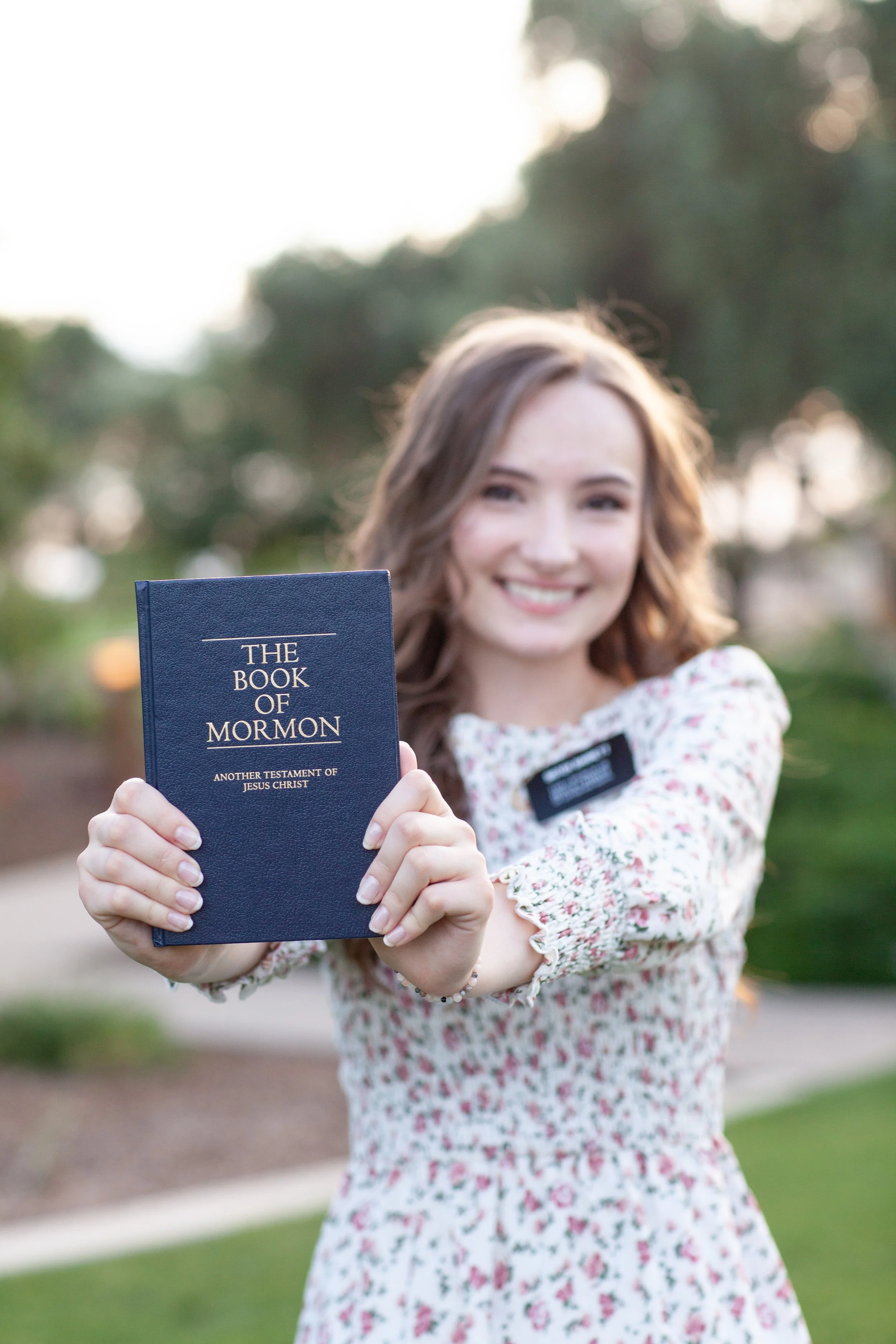 A young woman with wavy brown hair smiling while holding up a navy blue book titled "The Book of Mormon" outdoors in a park with trees in the background.