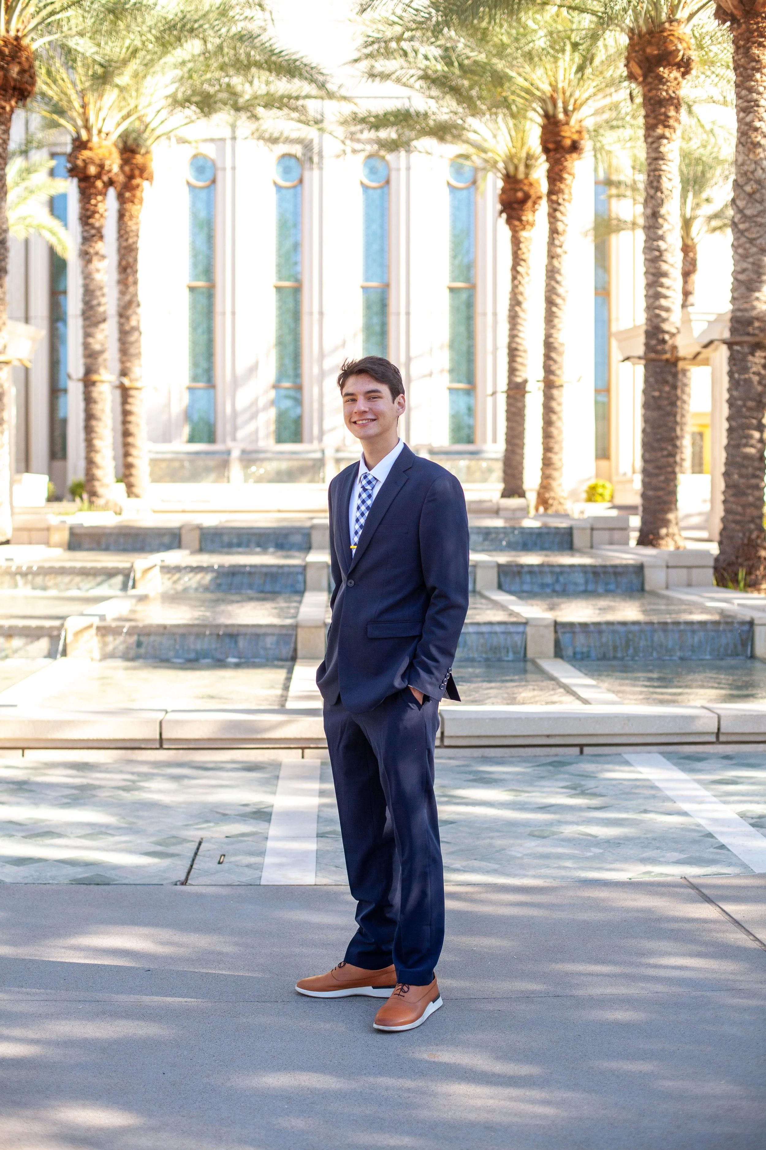 A young man in a navy blue suit and tan shoes standing outdoors in front of a modern building with tall windows and palm trees, smiling at the camera.