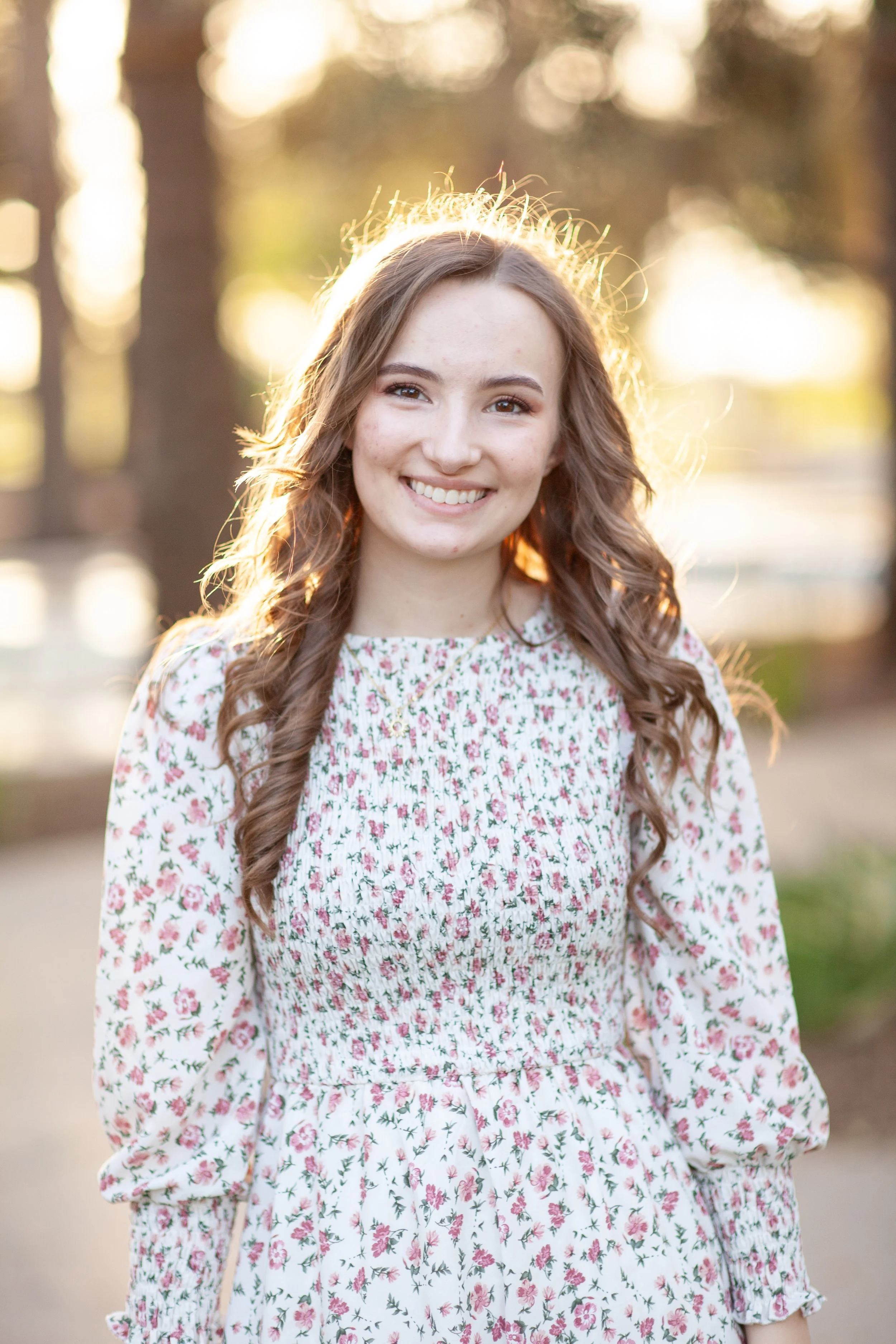 A young woman with long, wavy brown hair, smiling, standing outdoors during golden hour, with a background of trees and soft sunlight.