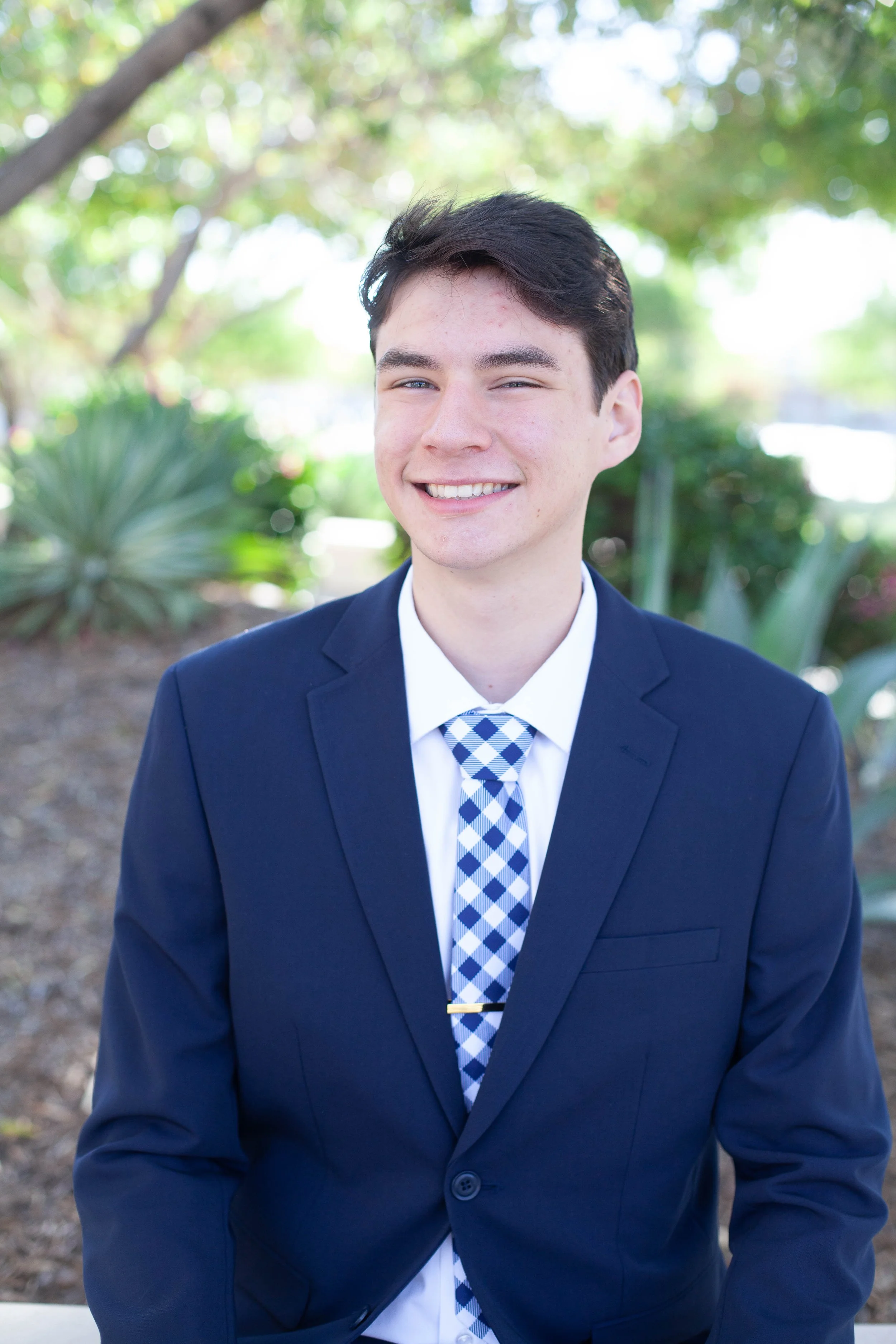 A young man with dark hair wearing a navy blue business suit, white shirt, and blue checkered tie, smiling outside with blurred green foliage in the background.