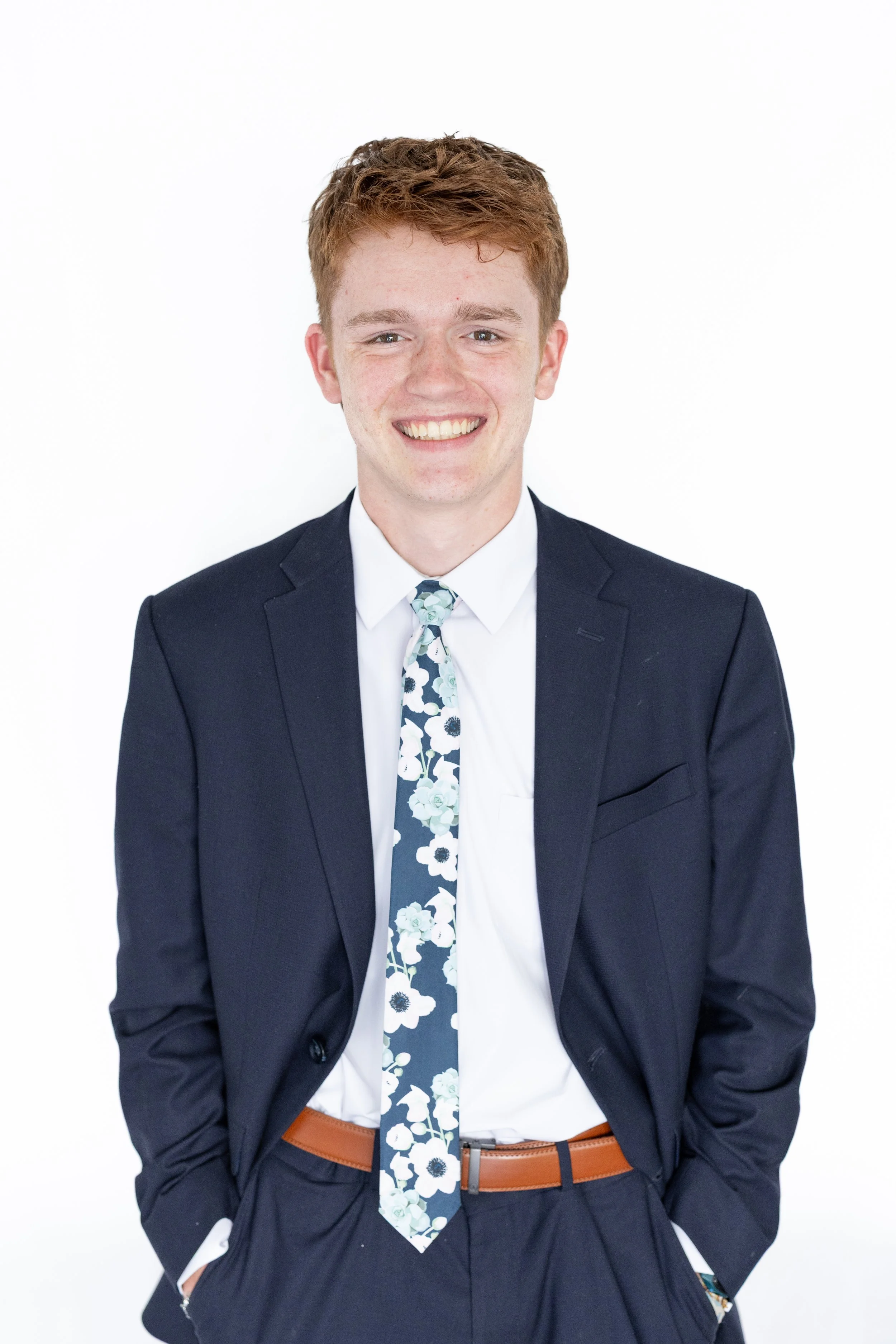 Young man with red hair in a suit smiling, wearing a white shirt and floral tie, standing against a plain white background.