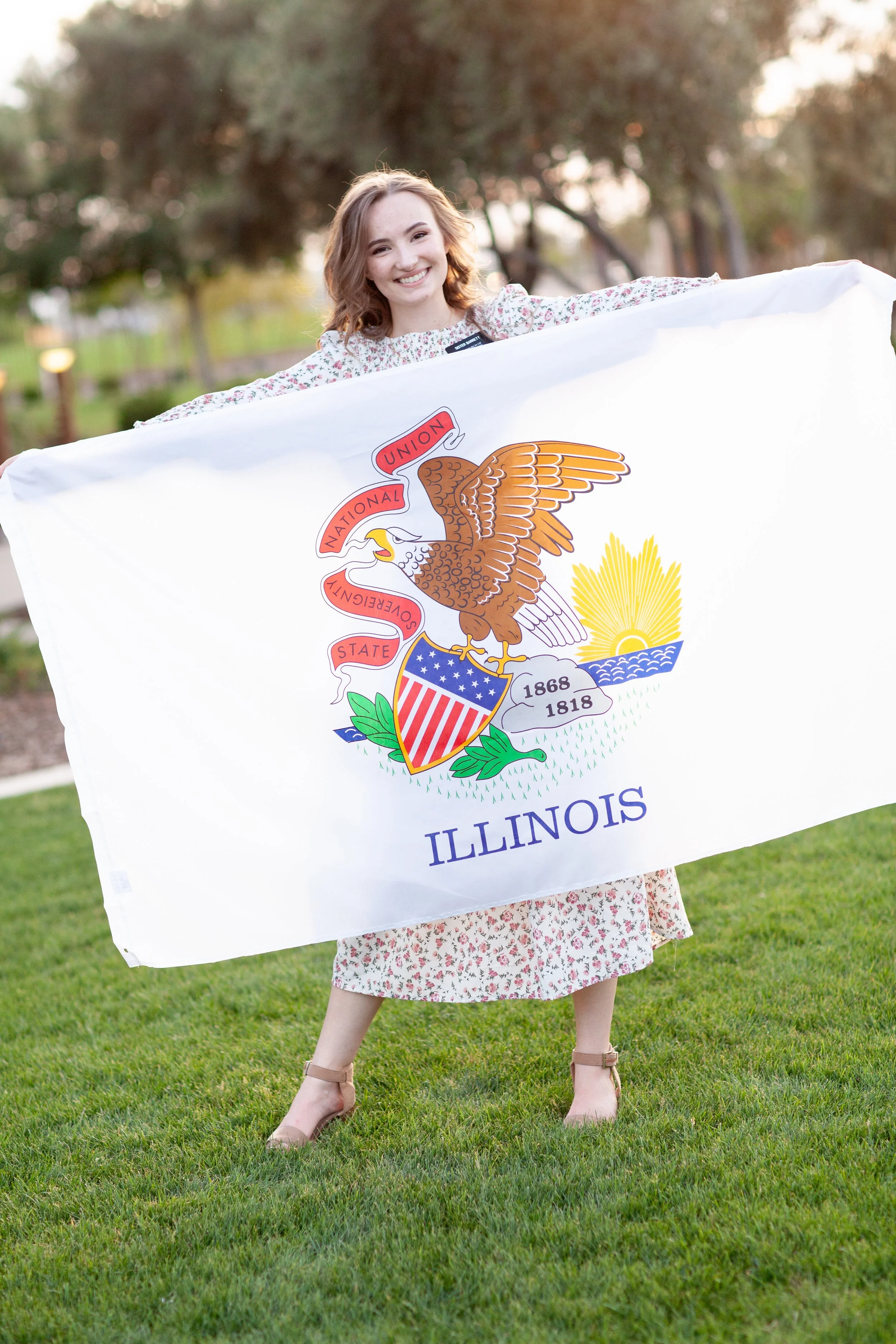 A girl holding a large Illinois state flag outdoors on grass, smiling, wearing a floral dress and nude heels.