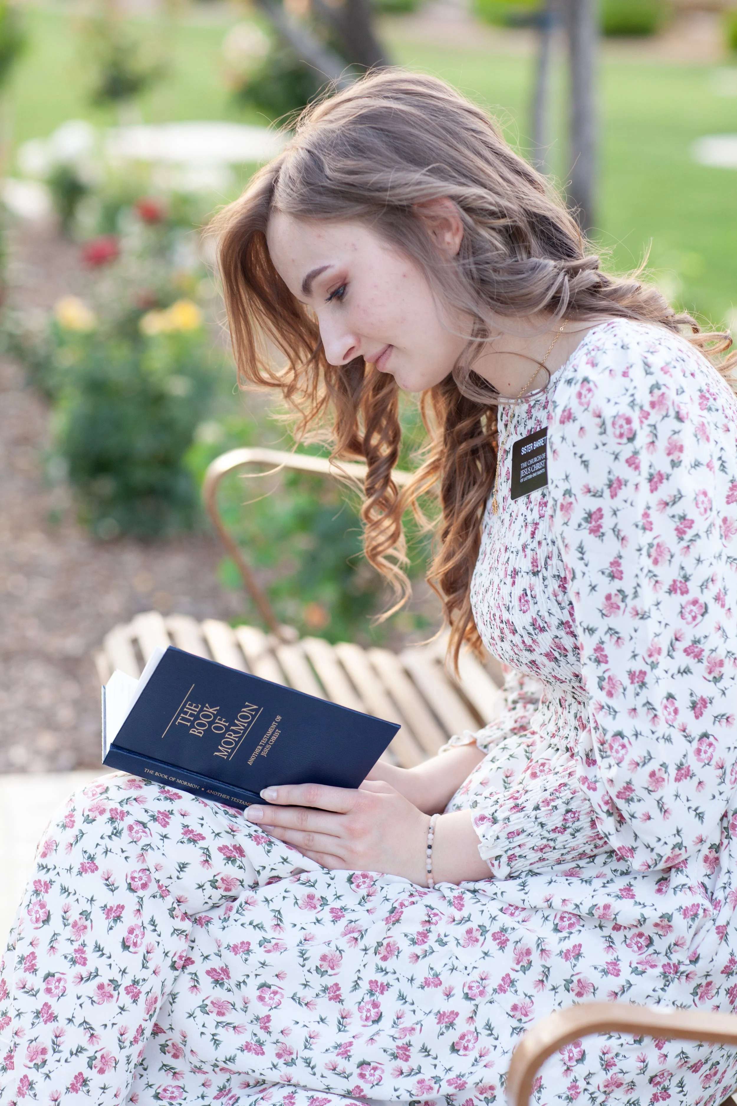 A young woman with long, wavy brown hair and light skin is sitting outdoors on a park bench, reading a dark blue book titled "The Book of Mormon". She is wearing a white dress with a pink and green floral pattern and a gold necklace. The background i