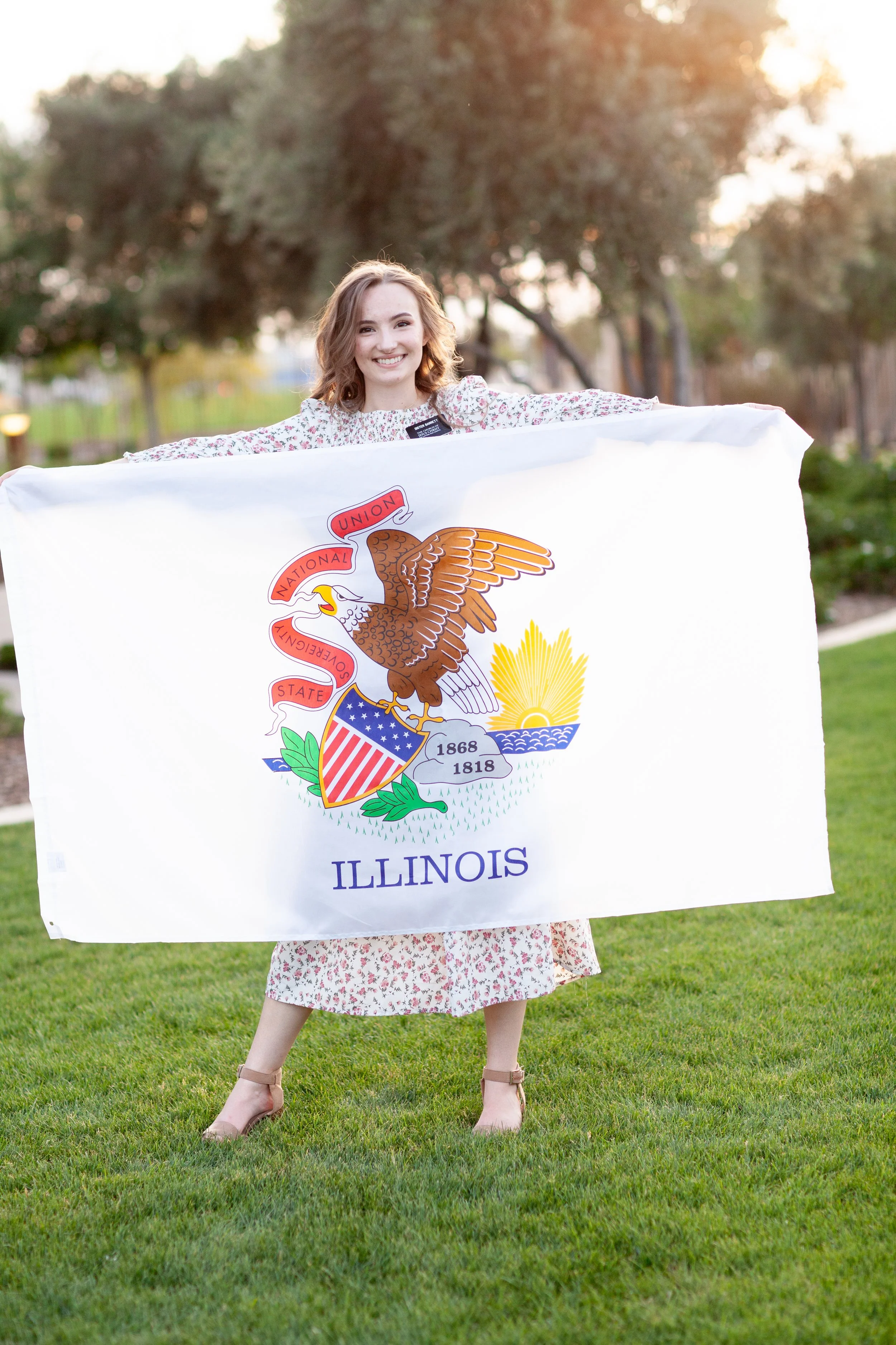 A young woman with curly, shoulder-length blonde hair is standing outdoors on a grassy area, holding up a large Illinois state flag that features the state seal with an eagle, shield, and sun, with the word "ILLINOIS" written below. She is smiling an