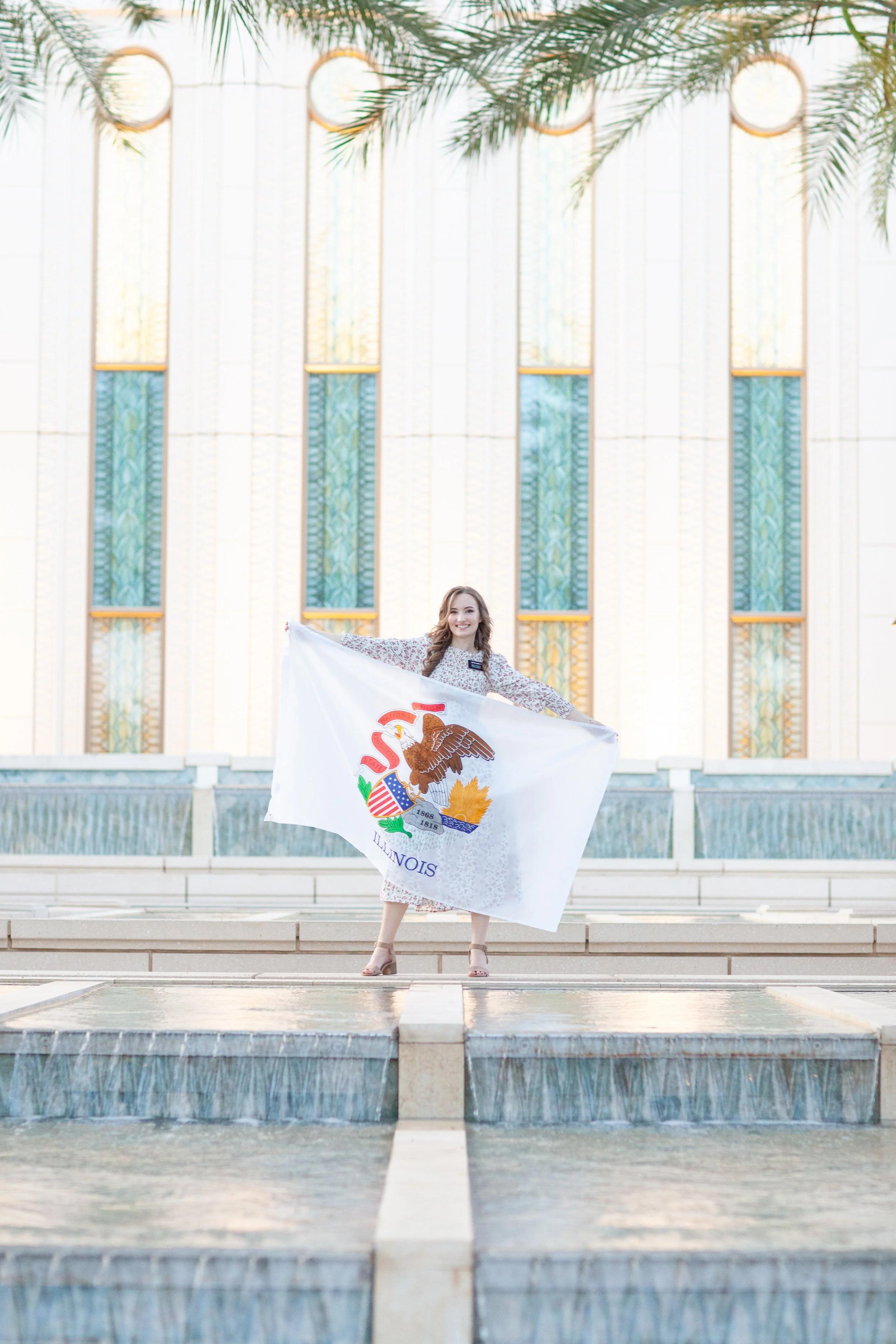 A young woman standing on steps in front of a building holding a large Illinois state flag. The building has large windows and palm tree branches above.