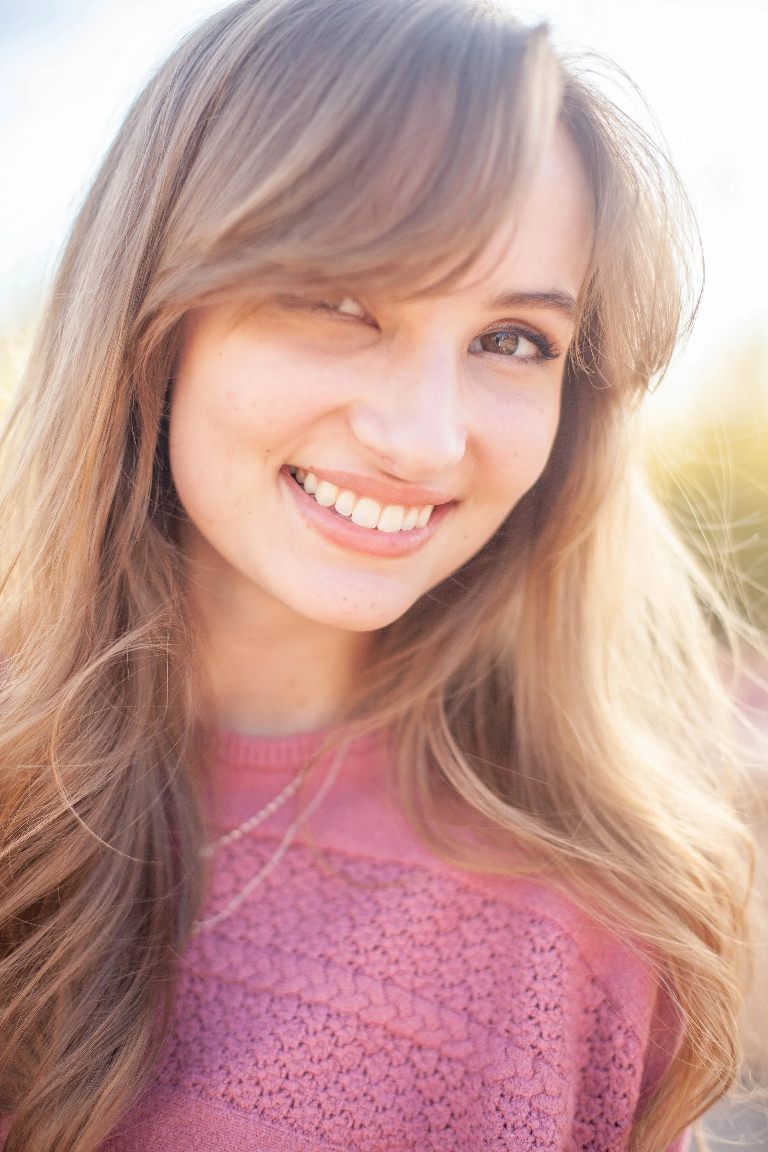 Close-up of a young woman smiling and winking, with long, wavy blonde hair, wearing a pink sweater and a necklace.