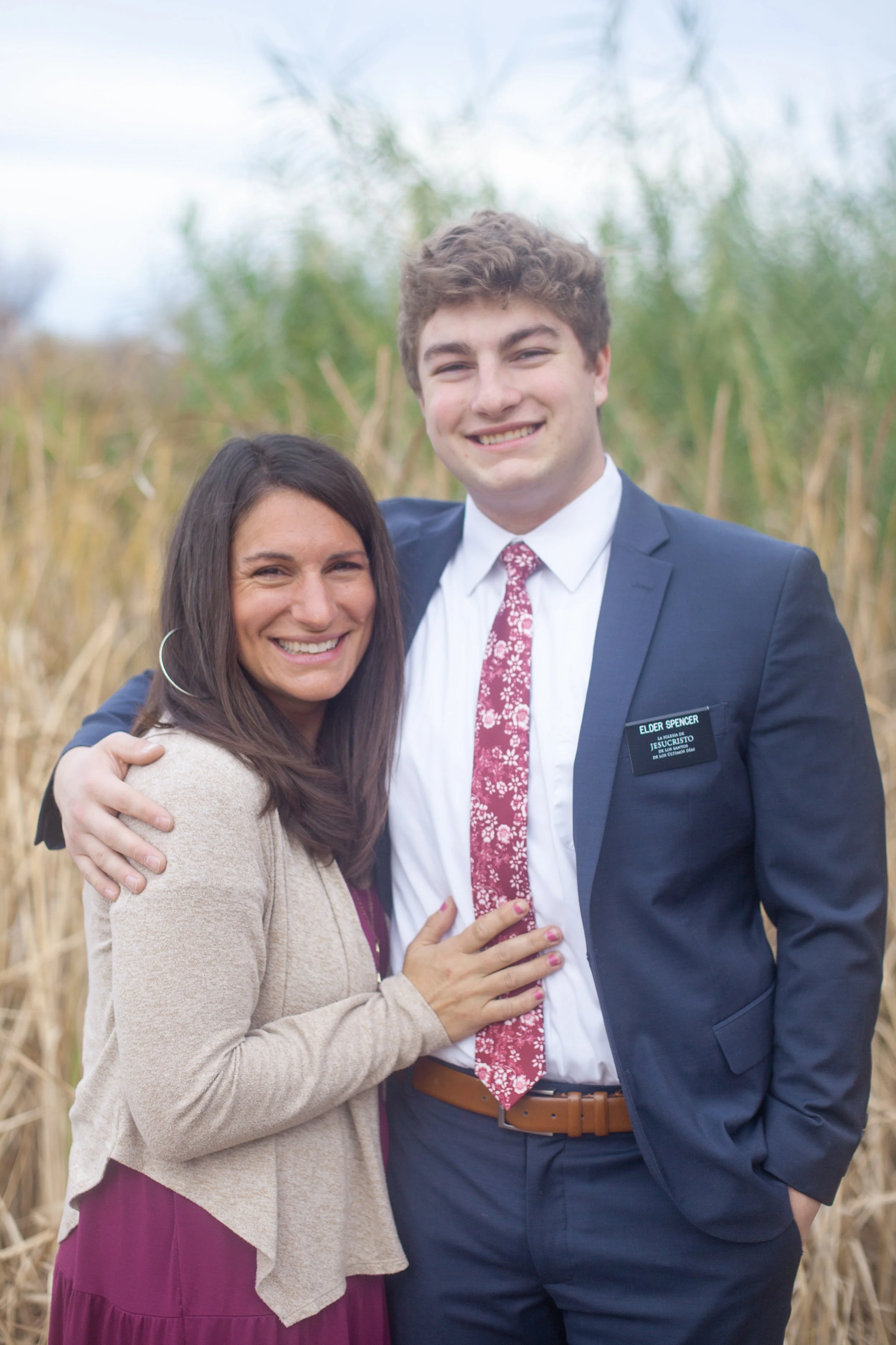 A woman and a man smiling and embracing outdoors with tall grass and trees in the background.