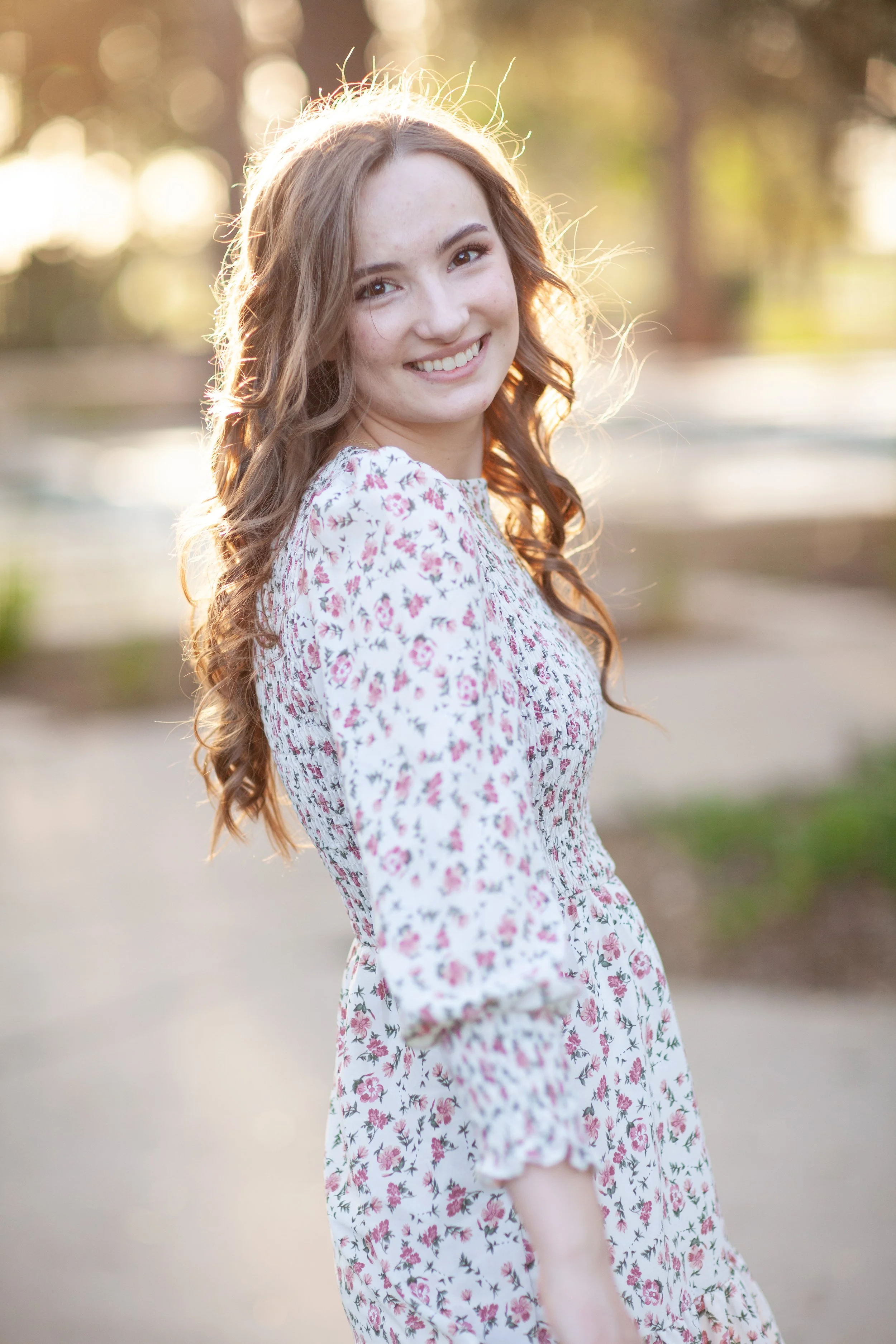 A young woman with long, curly brown hair smiling outdoors at sunset, wearing a floral dress with pink and white flowers.