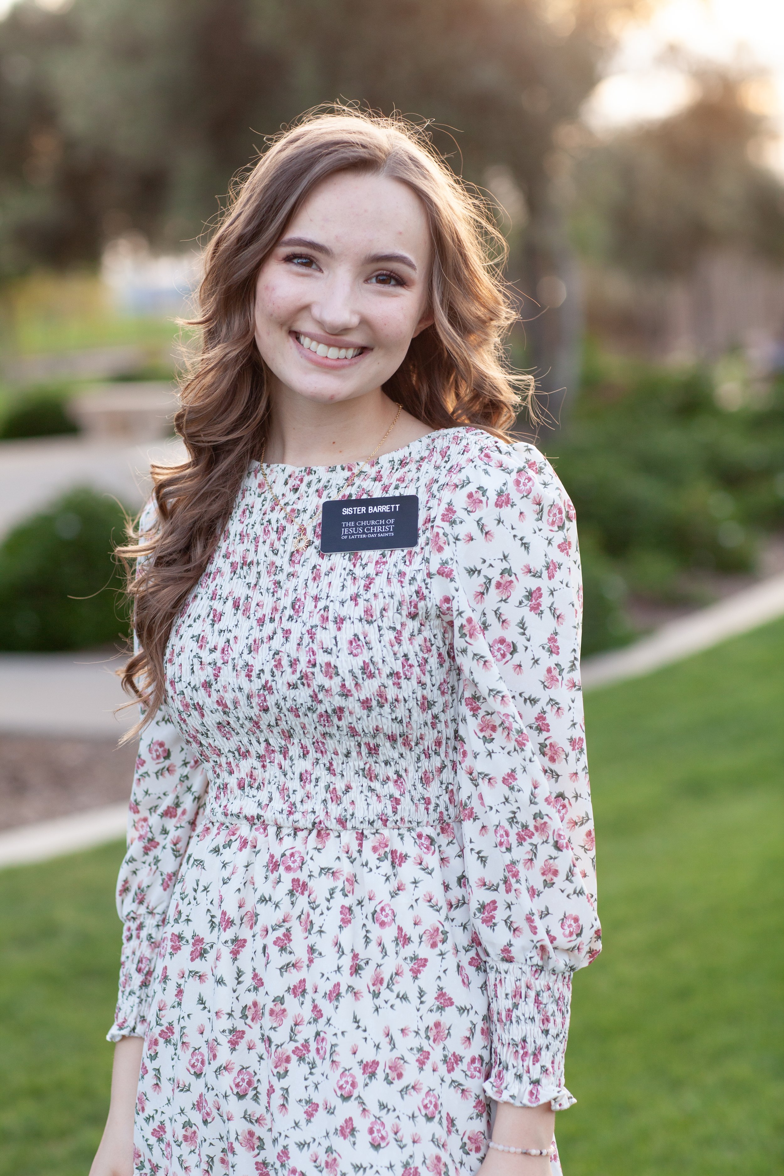 A young woman with long, wavy brown hair smiling outdoors during sunset, wearing a floral dress with a name tag that reads "SISTER BARRETT, THE CHURCH OF JESUS CHRIST OF LATTER-DAY SAINTS". She has a bracelet on her right wrist and is standing in a p