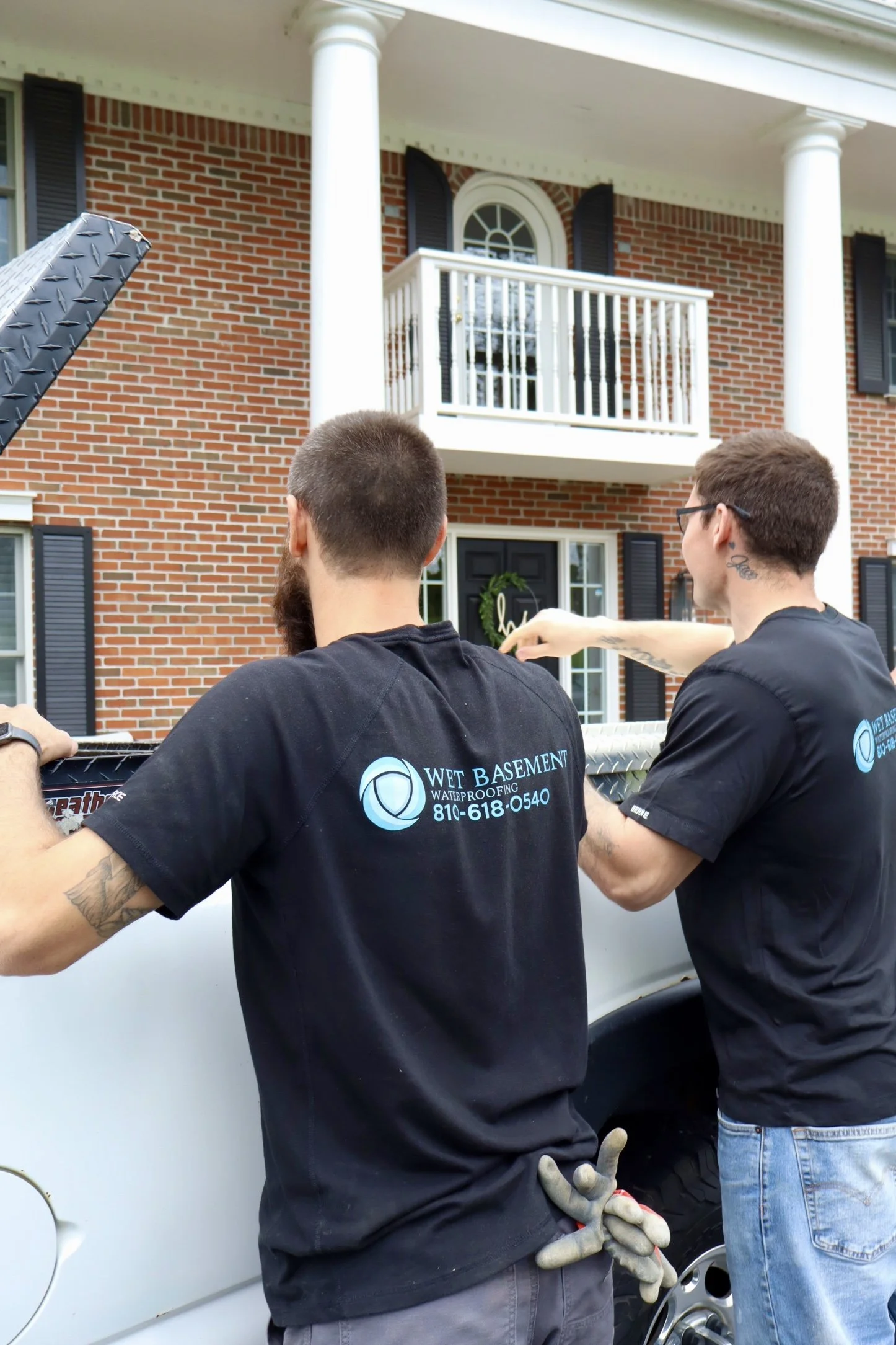 Two men working on a white truck during a roofing or construction project, with a brick house in the background, one of them holding gloves and wearing a black shirt with a waterproofing company logo.