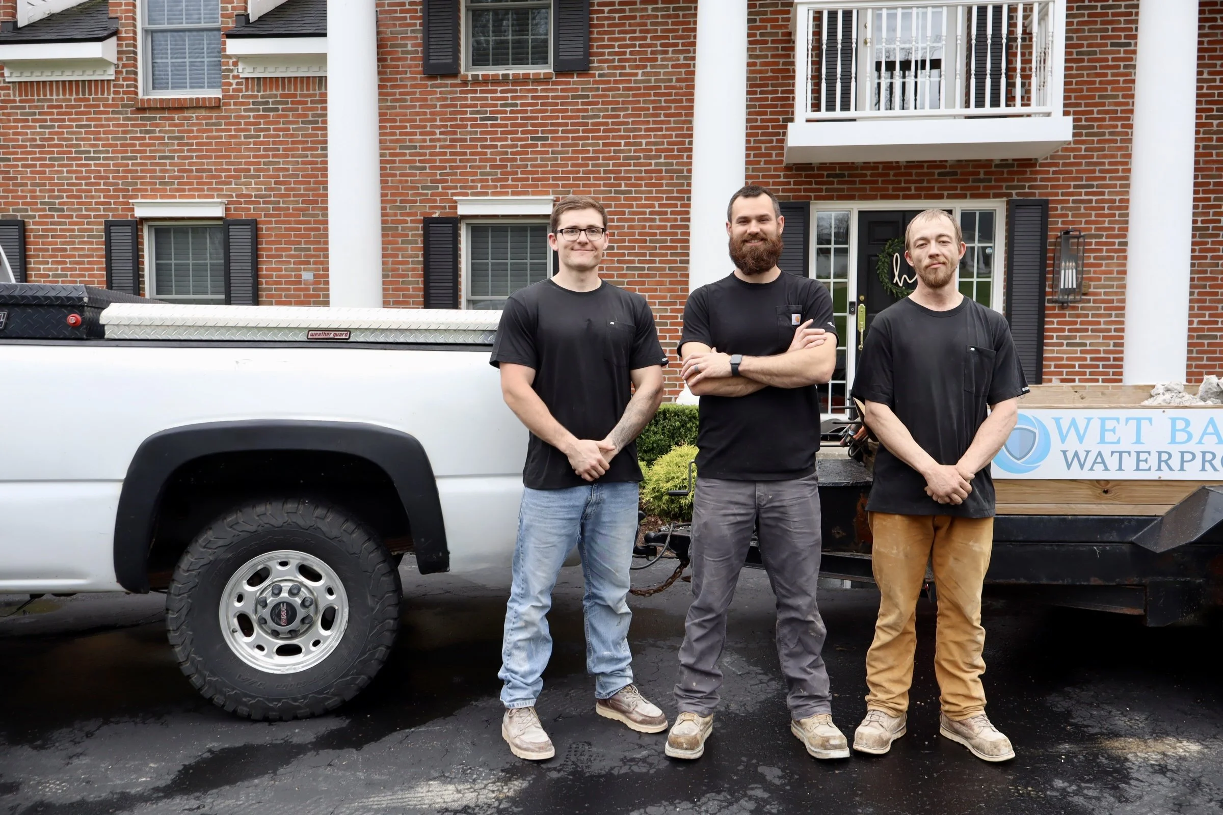Three men stand in front of a white pickup truck with a water delivery sign, parked outside a brick house with white columns and black shutters.