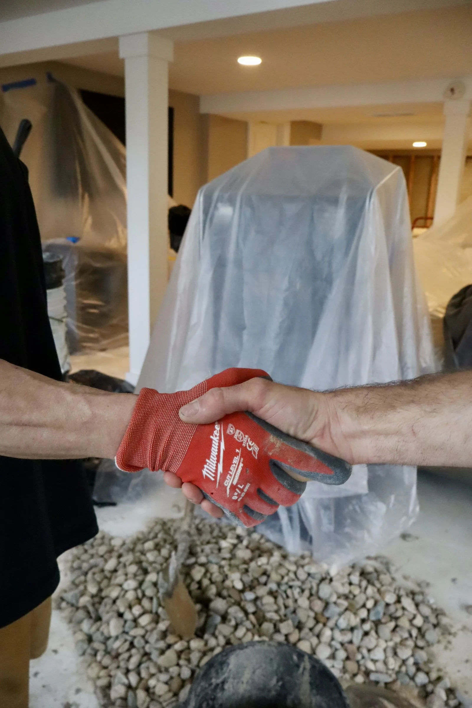 Two people shaking hands in a construction or renovation site, with one person's arm wearing a red glove with white text and the background showing covered objects and a rocky ground.