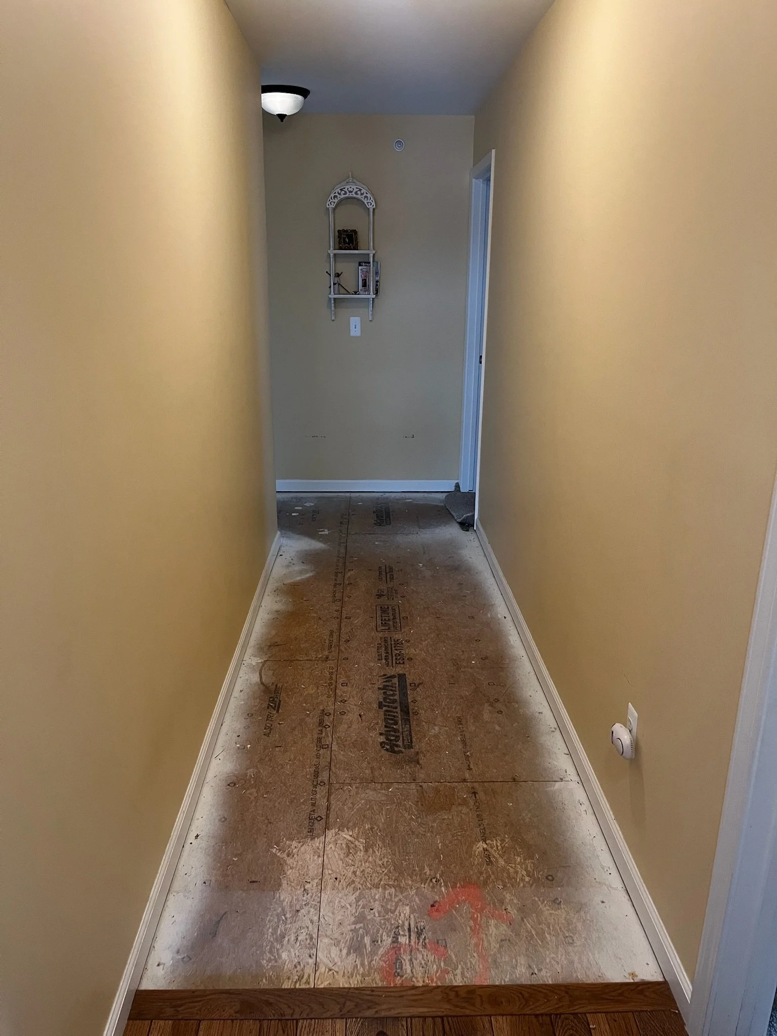 Empty hallway with yellow walls and uncarpeted floor under renovation, showing construction materials and a smoke detector on the ceiling.
