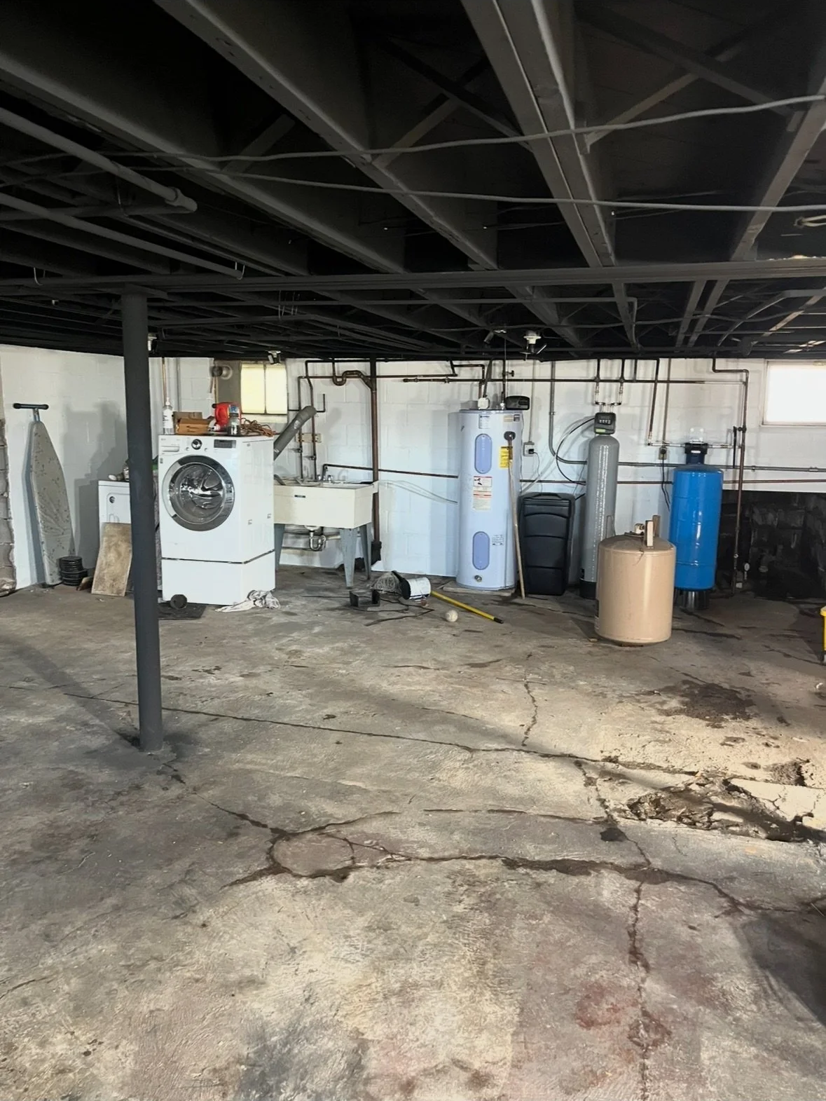 A cluttered basement laundry area featuring a washing machine, utility sink, water heater, and water treatment system, with a cracked concrete floor and exposed ceiling beams.