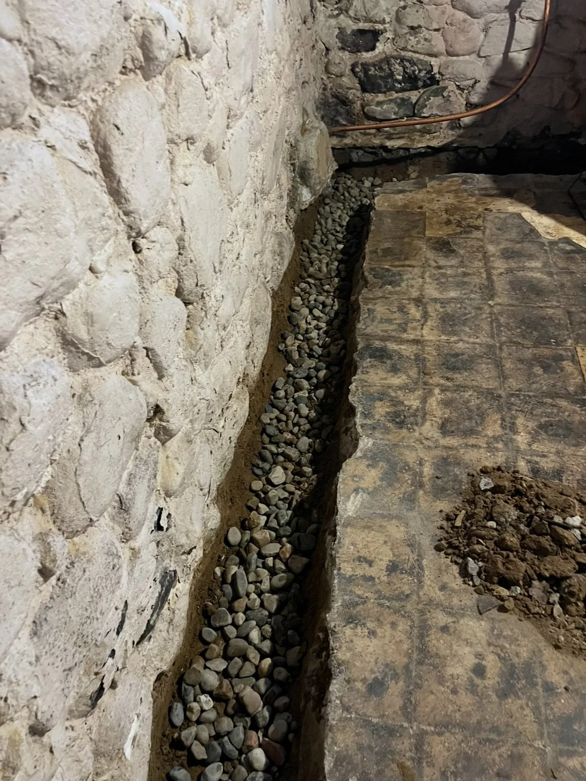 A stone wall with a trench filled with small gray rocks and earth, next to a worn floor made of wood or tiles, with some loose earth nearby, and a copper pipe in the corner.